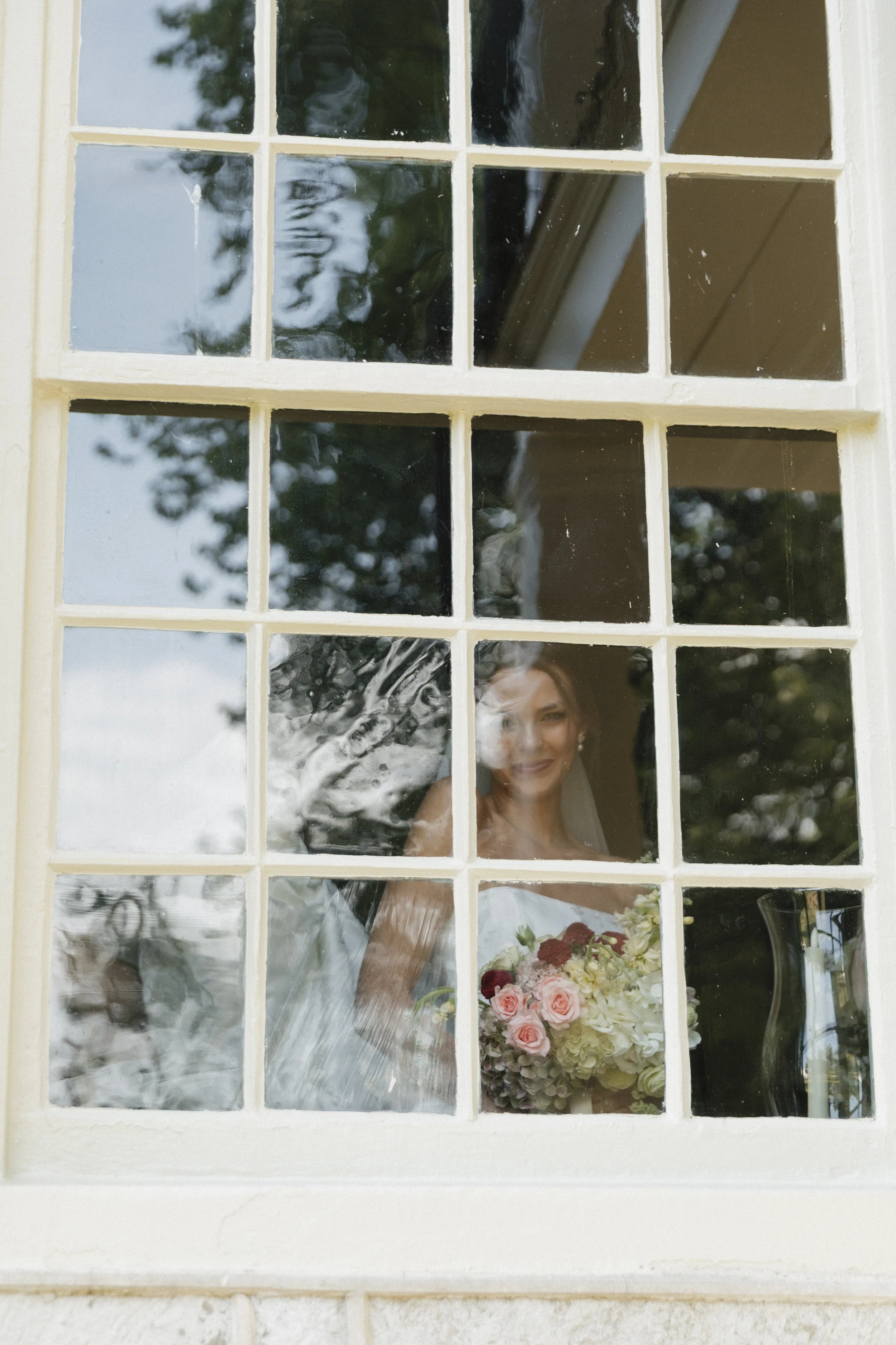 A woman in a wedding dress looking through a grid of glass panes on a window, with a bouquet of flowers in front of her, in an outdoor setting.