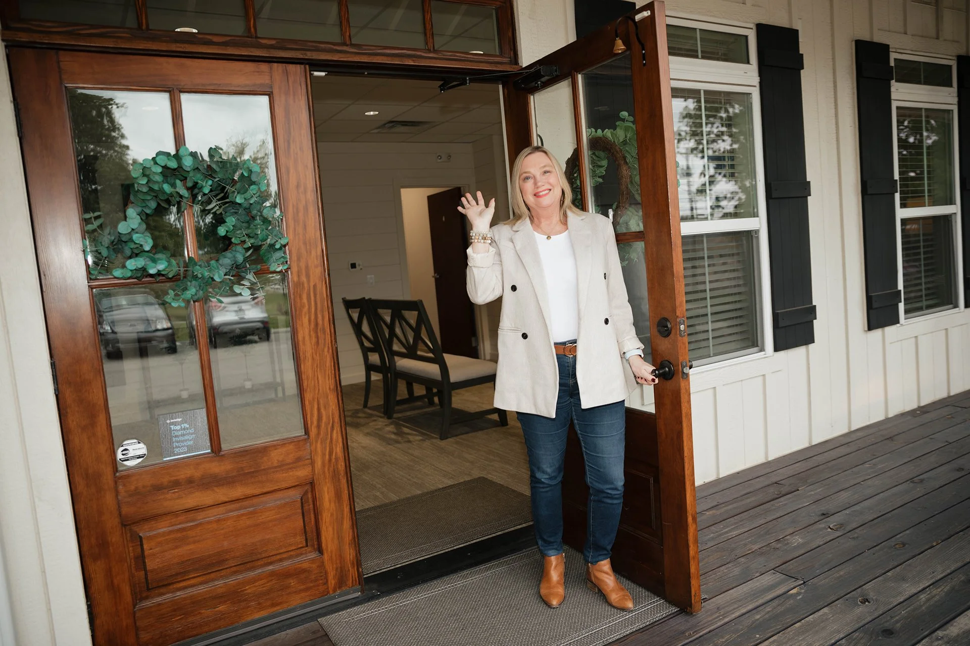 Woman standing at the porch door of a house, smiling and waving, dressed in a beige blazer, white top, blue jeans, and brown boots, with a decorated wreath hanging on the door.