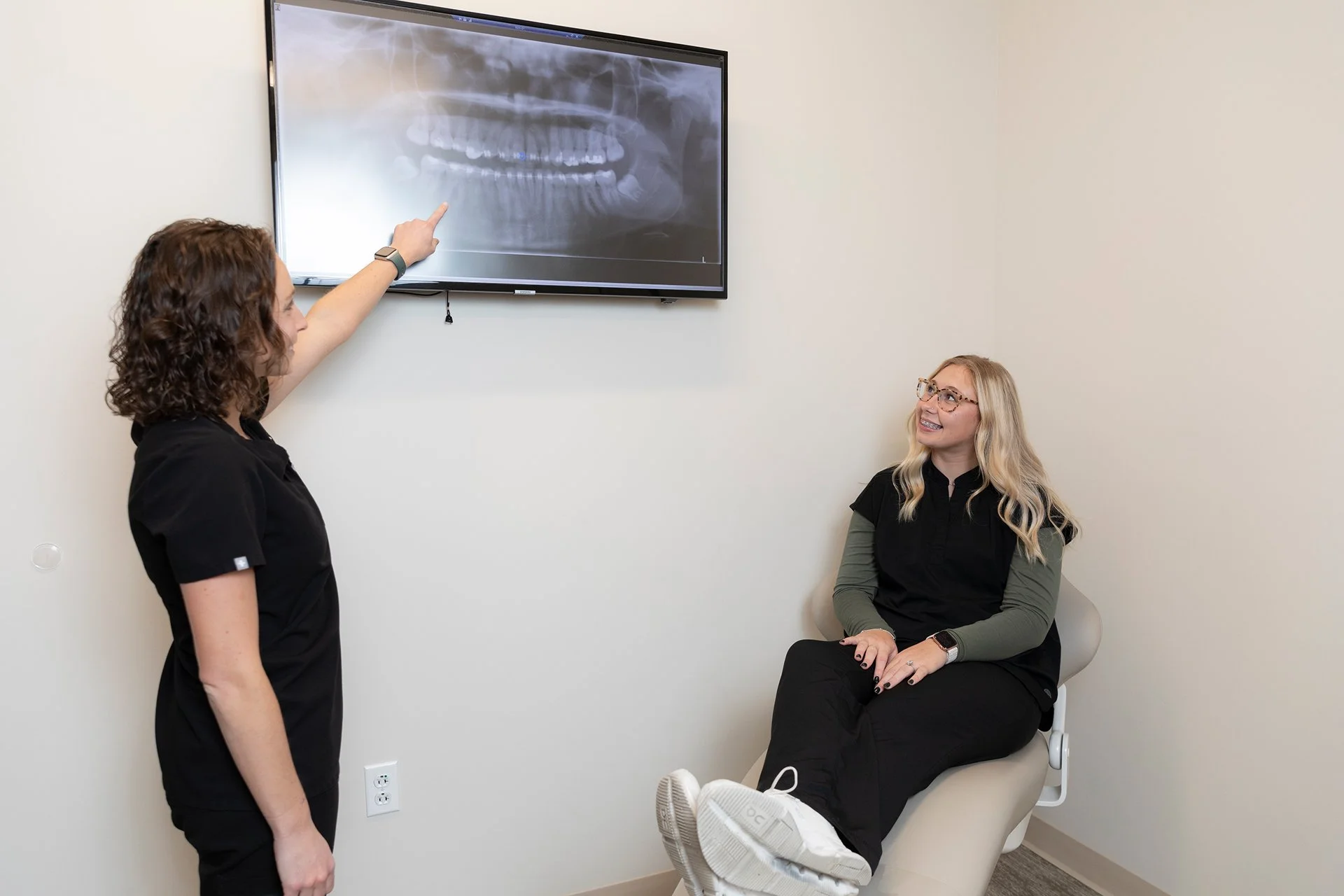 A healthcare professional pointing at a dental X-ray displayed on a wall monitor while a patient, sitting in a chair, listens and smiles in a medical office.