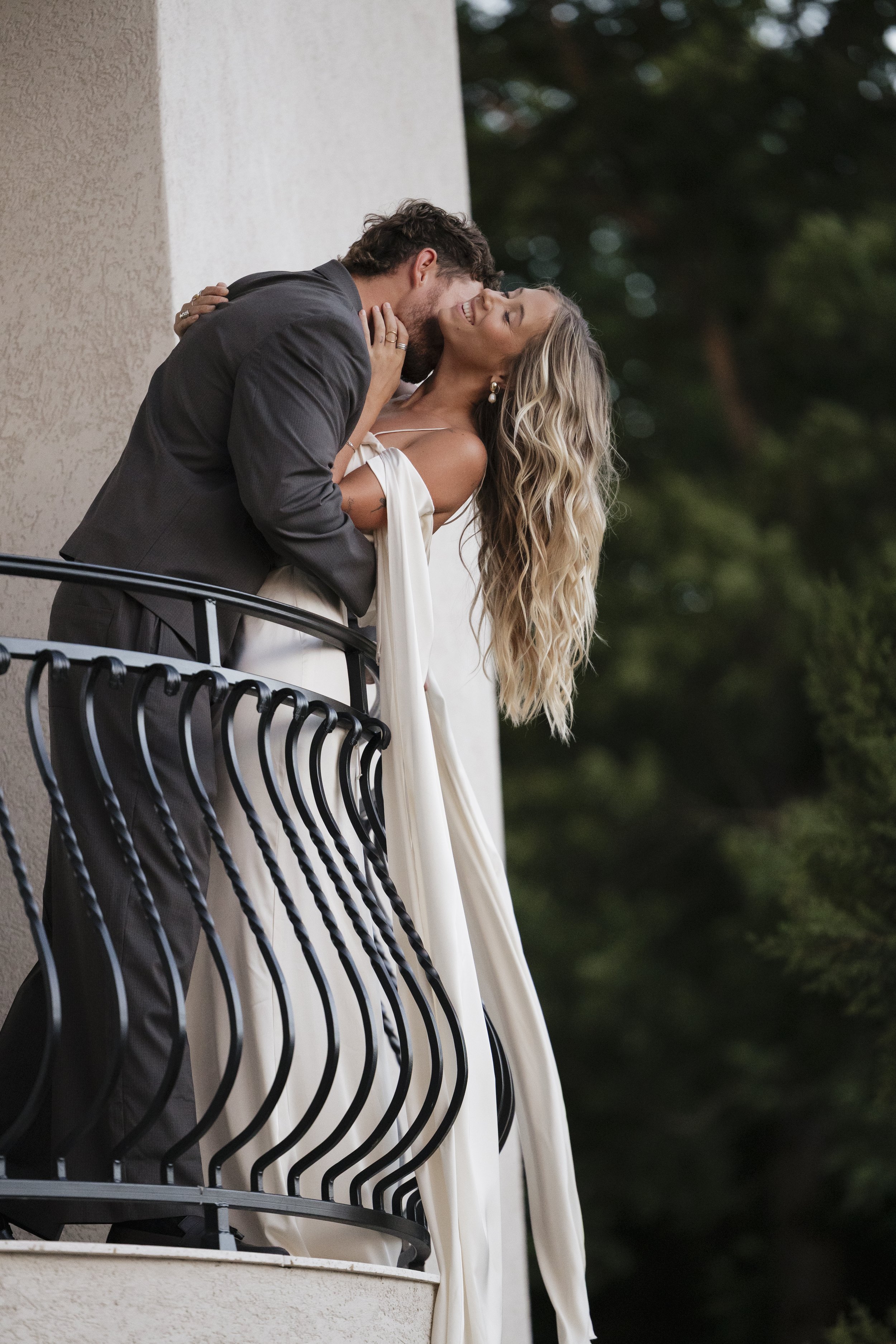 A man and woman are on a balcony, sharing a kiss. The woman has long, wavy blonde hair and is wearing a white dress. The man has dark hair and is dressed in a dark suit. They are smiling and holding each other, with a blurred background of trees.