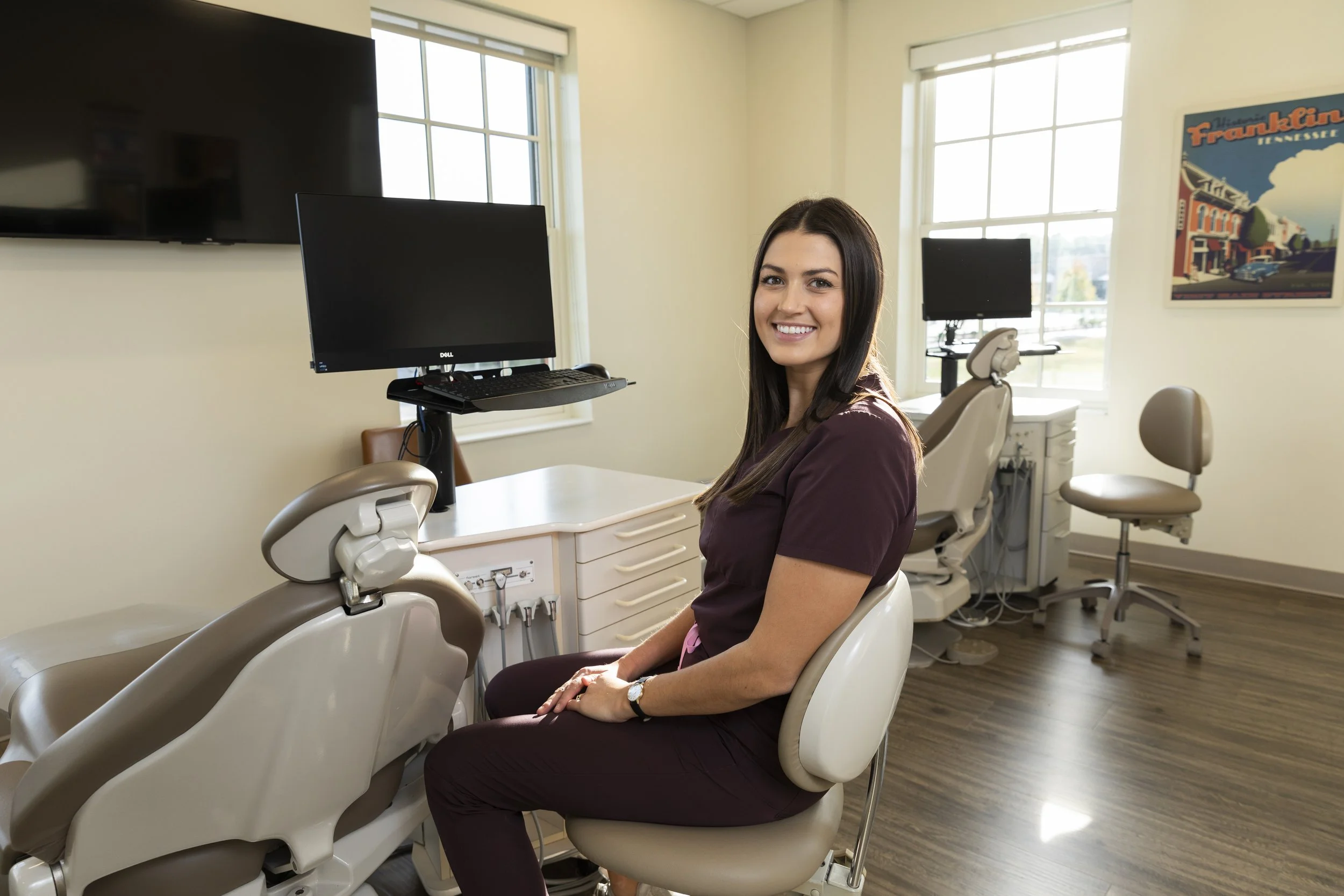 A woman sitting in a dental clinic's dental chair, smiling, with dental equipment and monitors in the background.