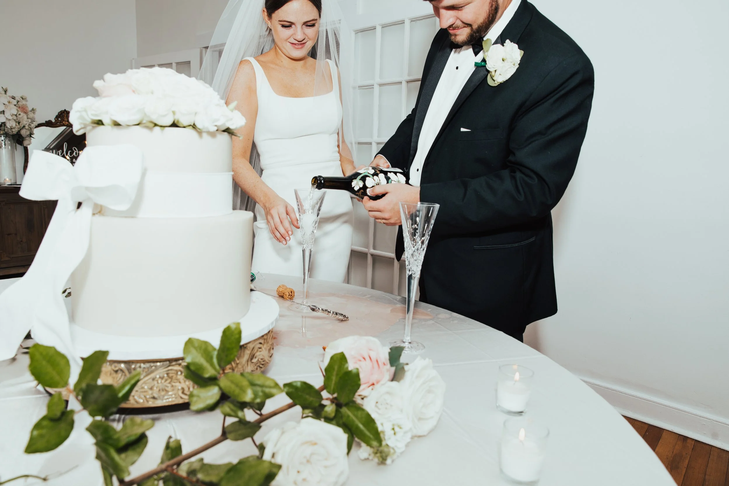 Bride and groom pouring champagne into glasses at their wedding reception, with a wedding cake and floral decorations in the foreground.