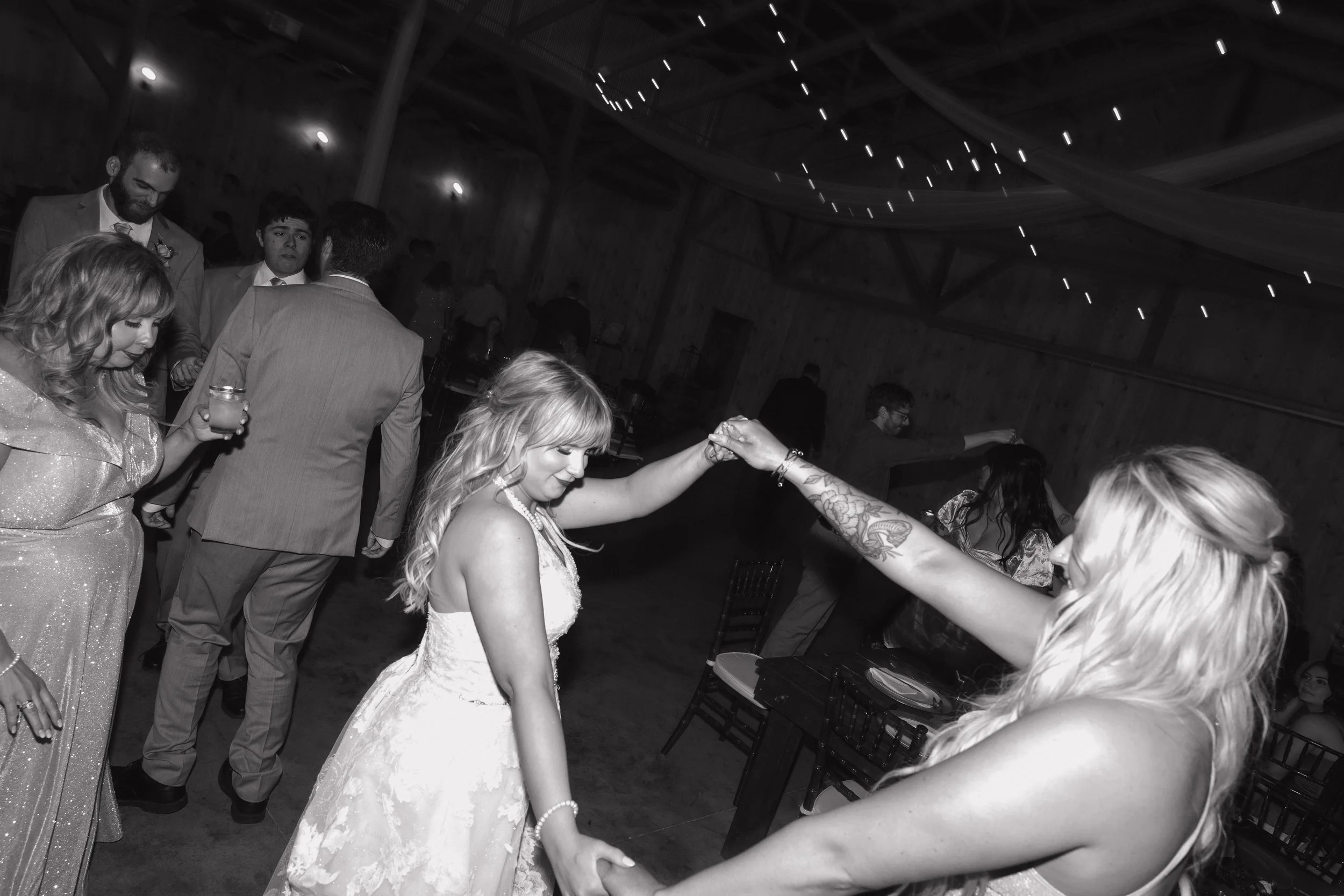 People dancing at a wedding reception in a rustic barn, with string lights on the ceiling and wooden walls.