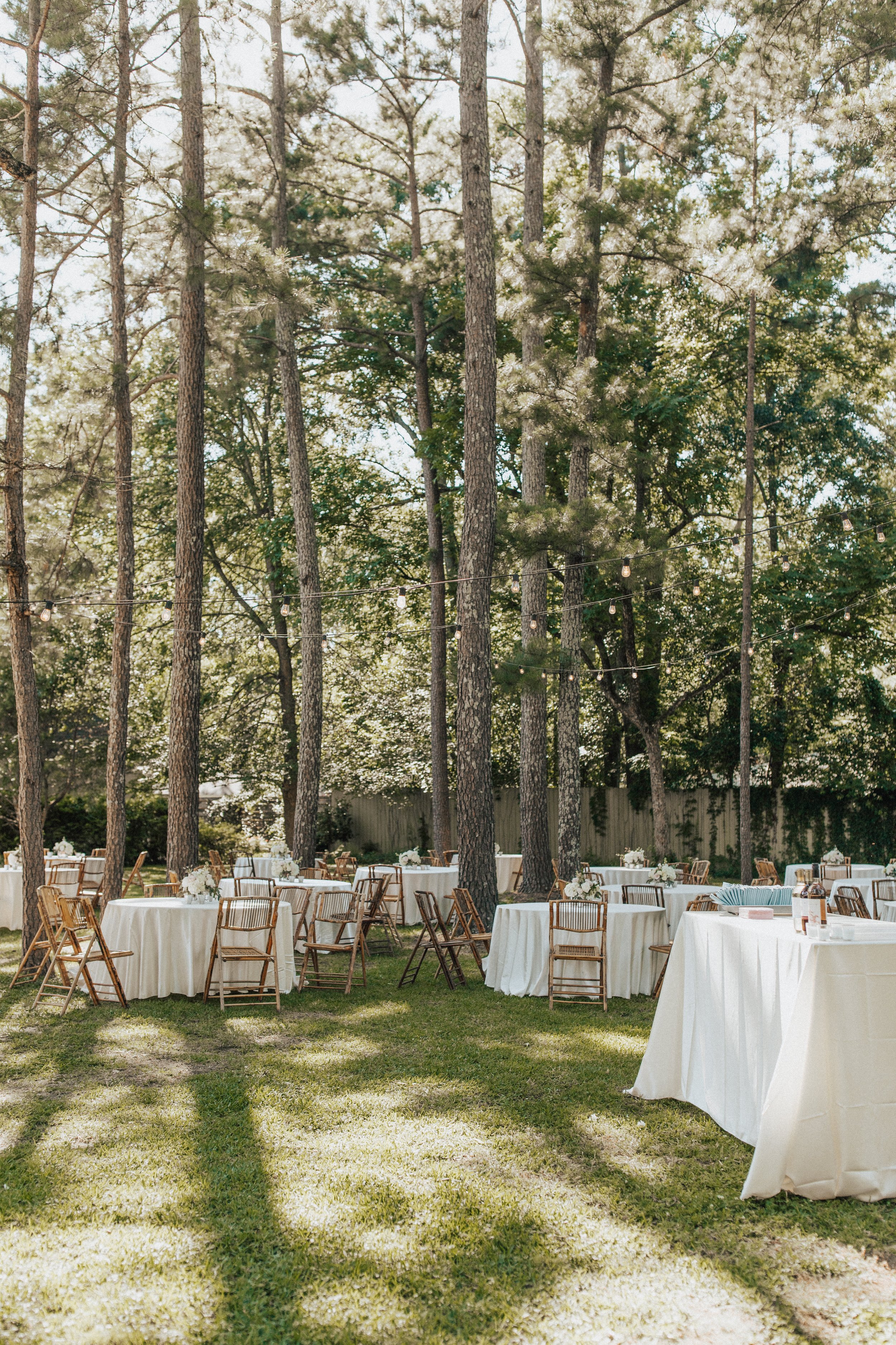 Outdoor wedding reception setup with round tables covered in white tablecloths, wooden chairs, floral centerpieces, surrounded by tall trees, with string lights hanging above on a sunny day.