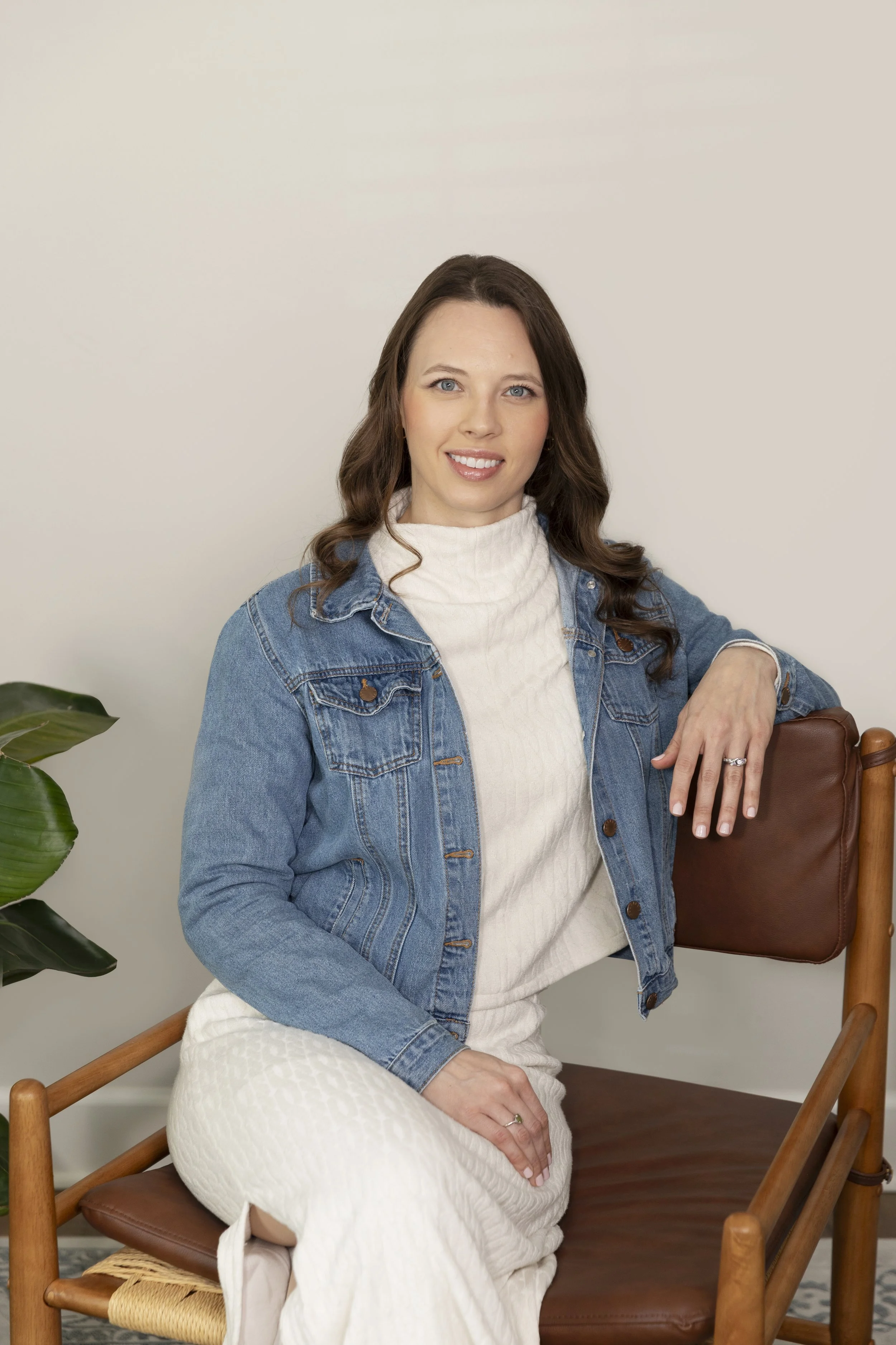 A woman with long brown hair sitting on a wooden chair with a brown leather backrest, wearing a white turtleneck and a denim jacket, against a plain off-white background.