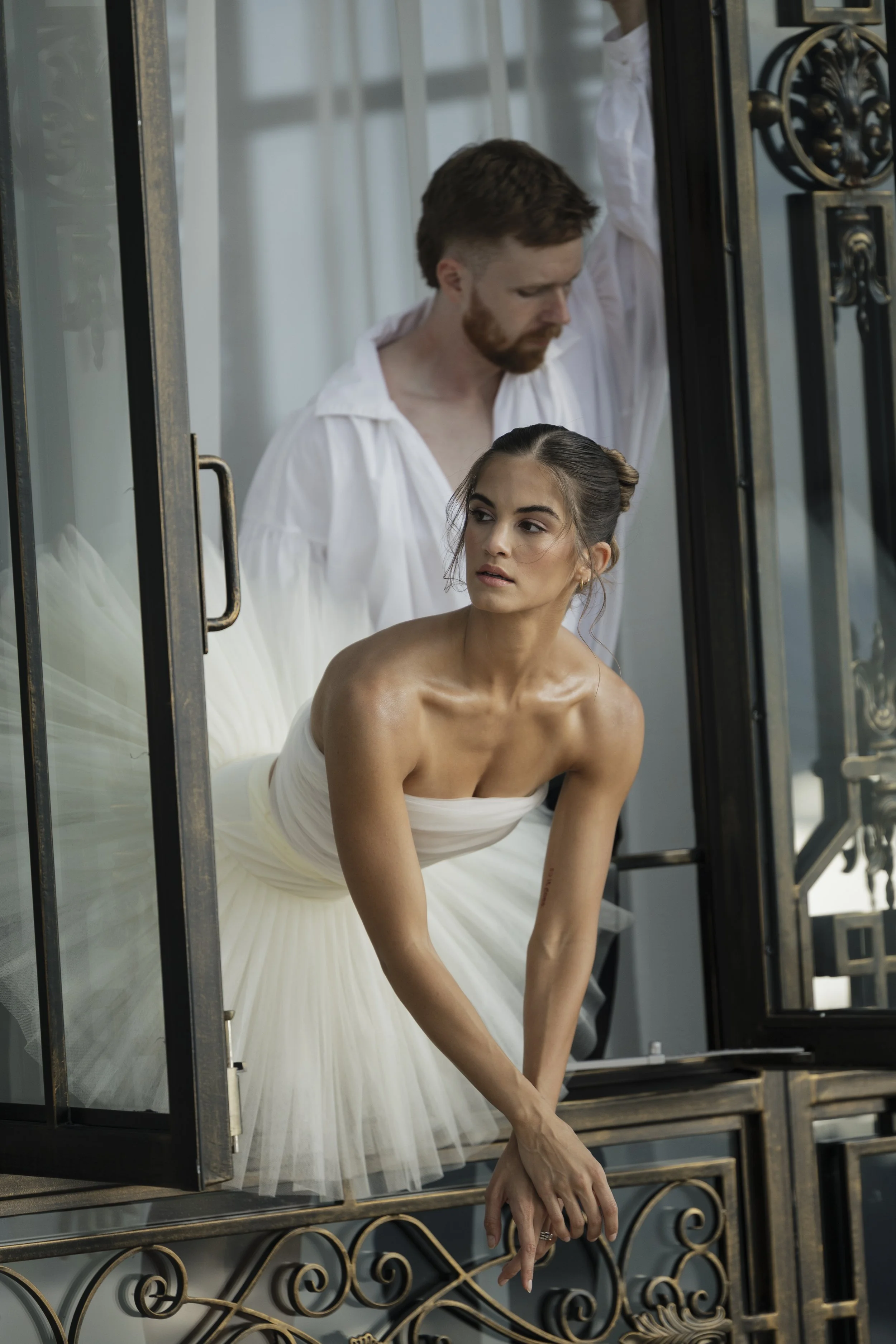 A woman in a white dress leaning out of a window with an ornate black metal frame, while a man in a white shirt stands behind her.