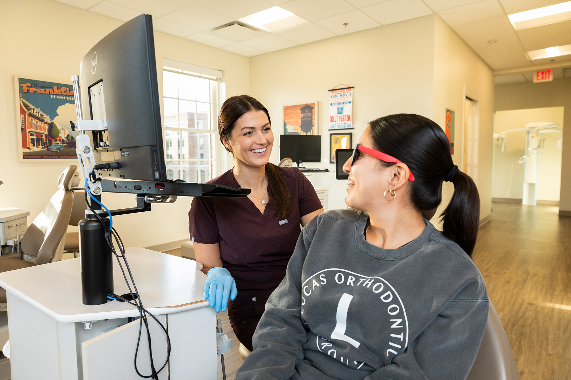 A female healthcare professional smiling at a female patient at a medical clinic. The patient, wearing glasses and a gray sweatshirt, is seated facing the healthcare worker. The scene is well-lit with posters on the wall behind, and a computer monitor to the side.