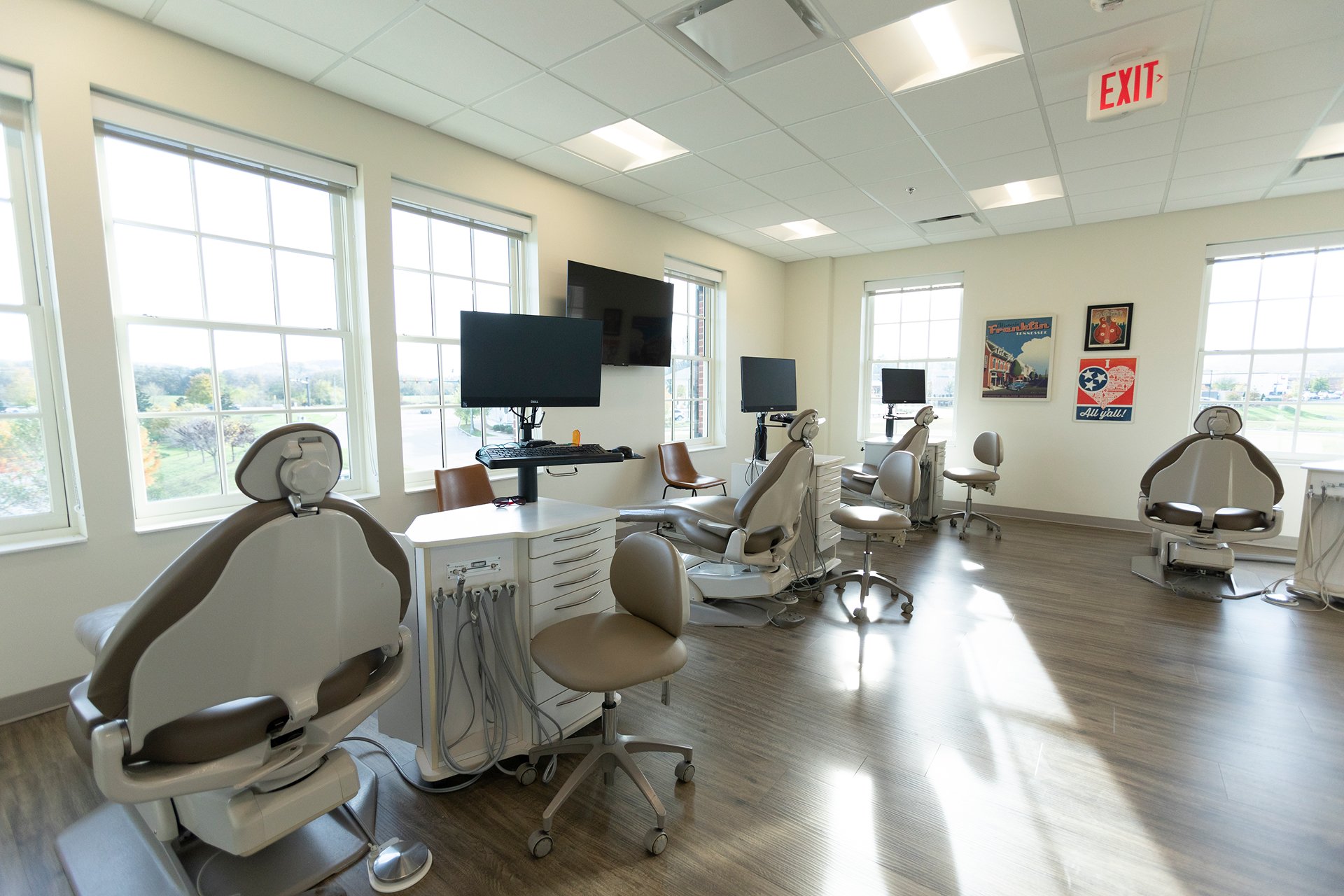 Dental treatment room with chairs, computers, and medical equipment, illuminated by natural light from large windows.