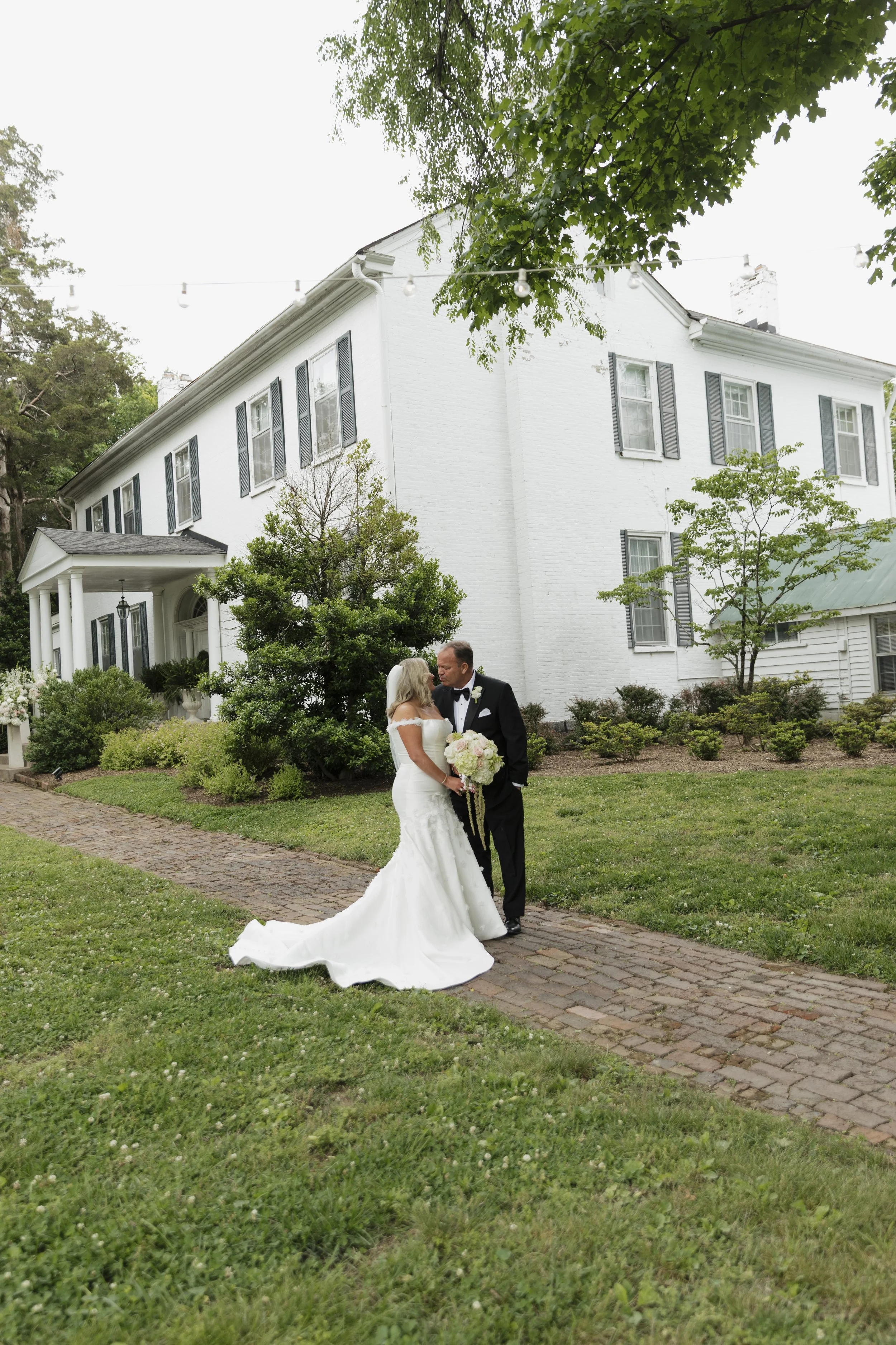 A bride and groom share a kiss outdoors in front of a large white house with dark shutters, surrounded by greenery and trees, during their wedding ceremony.