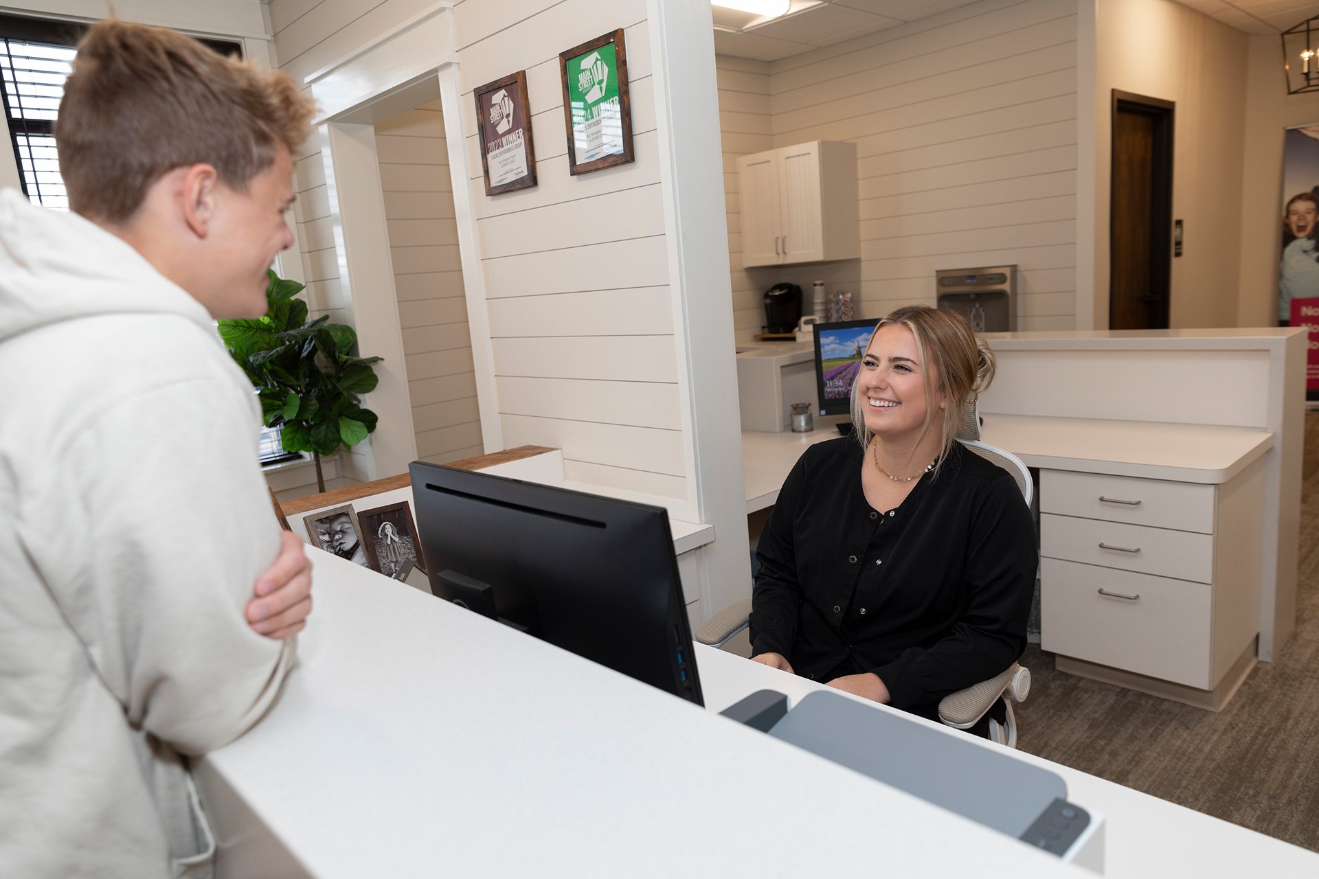 A smiling woman sitting at a reception desk talking to a young man who is standing. The woman has blonde hair and is wearing a black shirt. The man has light brown hair and is wearing a white hoodie. The reception area has white shiplap walls, framed awards, a computer, and decorative photographs on the desk. There is a large leafy green plant near the window and a woman in the background behind a doorway, smiling.