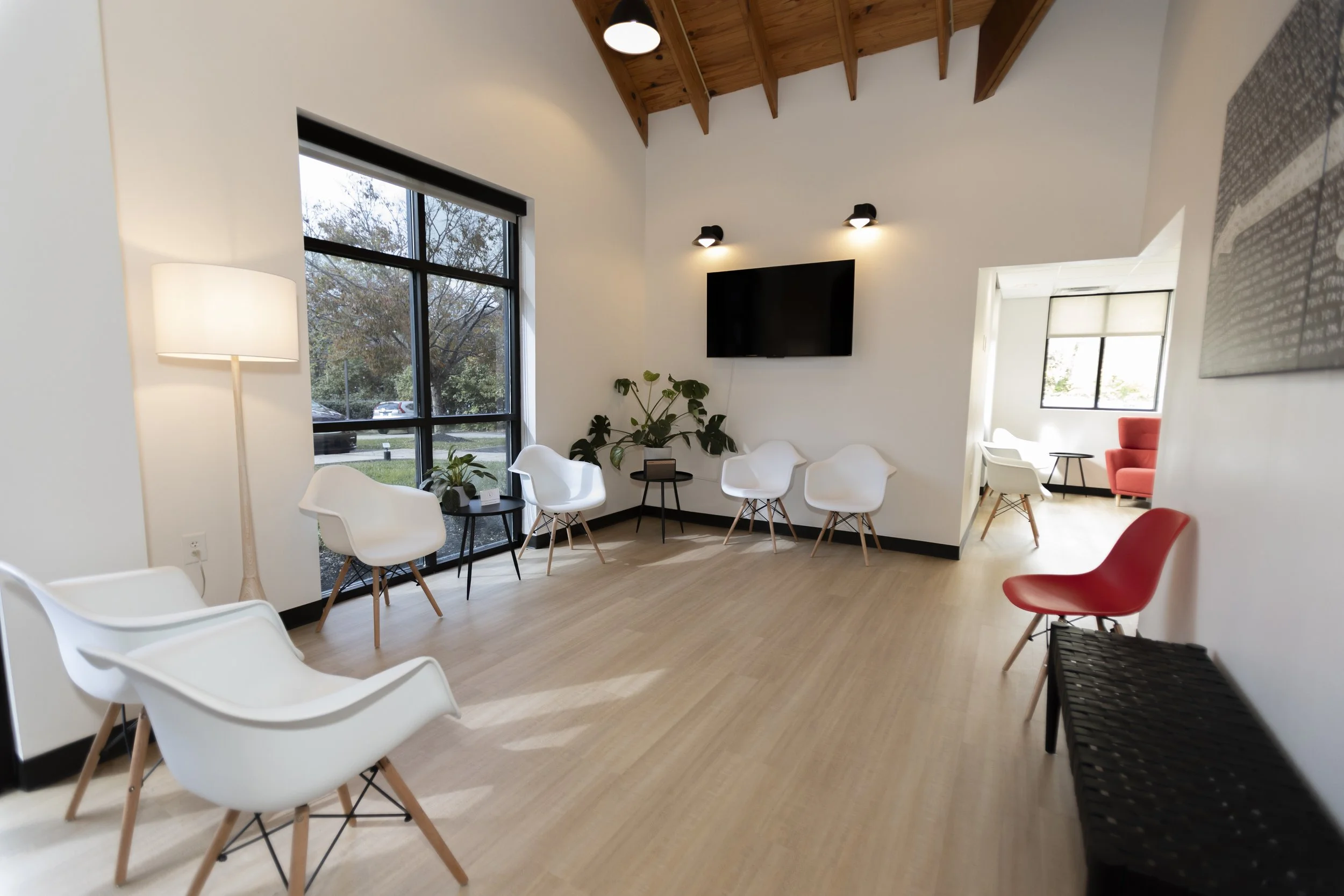 Modern waiting room with white chairs, a red chair, a black bench, a tall white lamp, a large window, green plants, a wall-mounted TV, and a separate room with a bright red armchair.