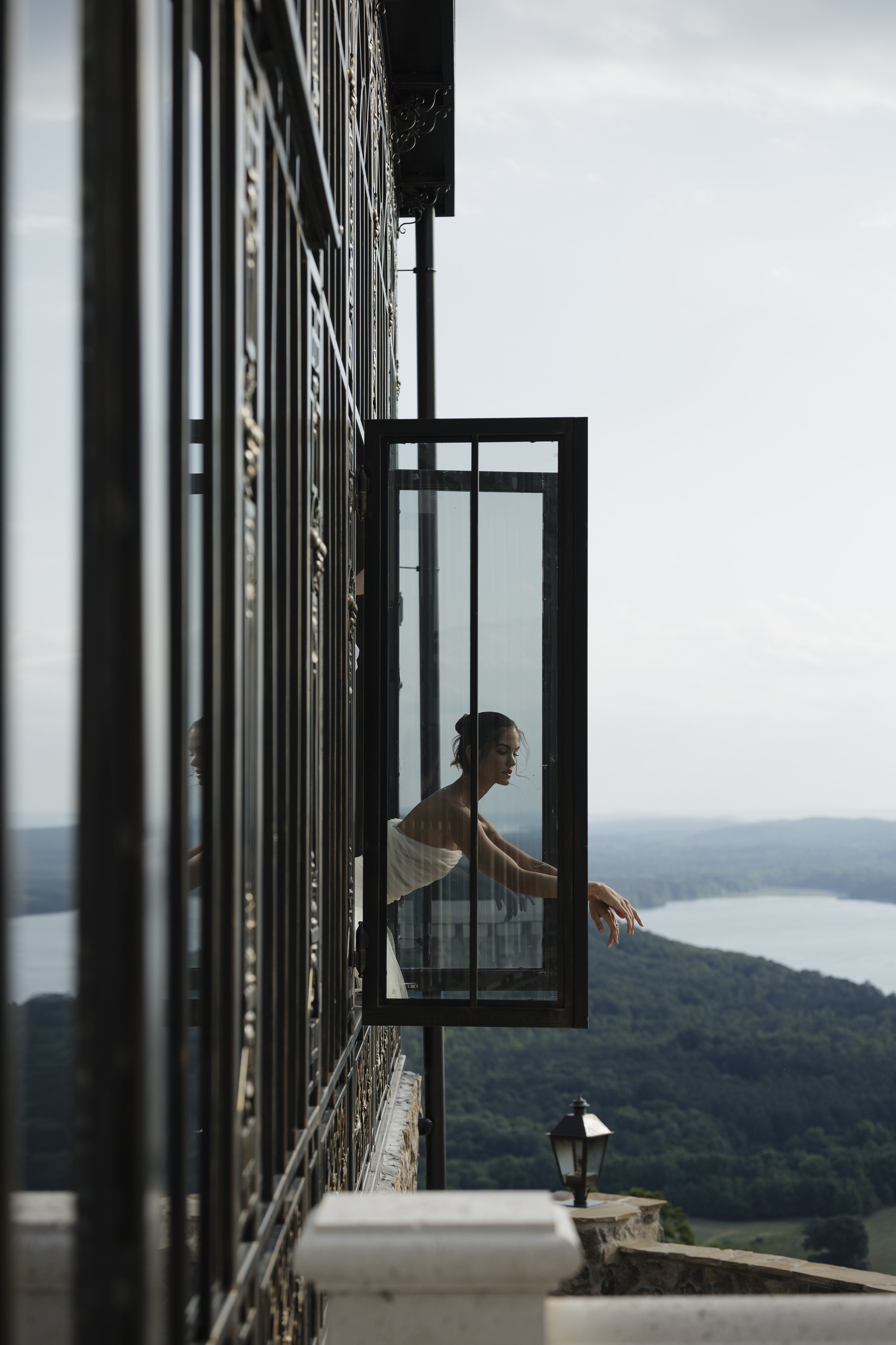 A woman in a strapless white dress standing at an open window on a building, overlooking a landscape with trees and a large body of water, during daytime.