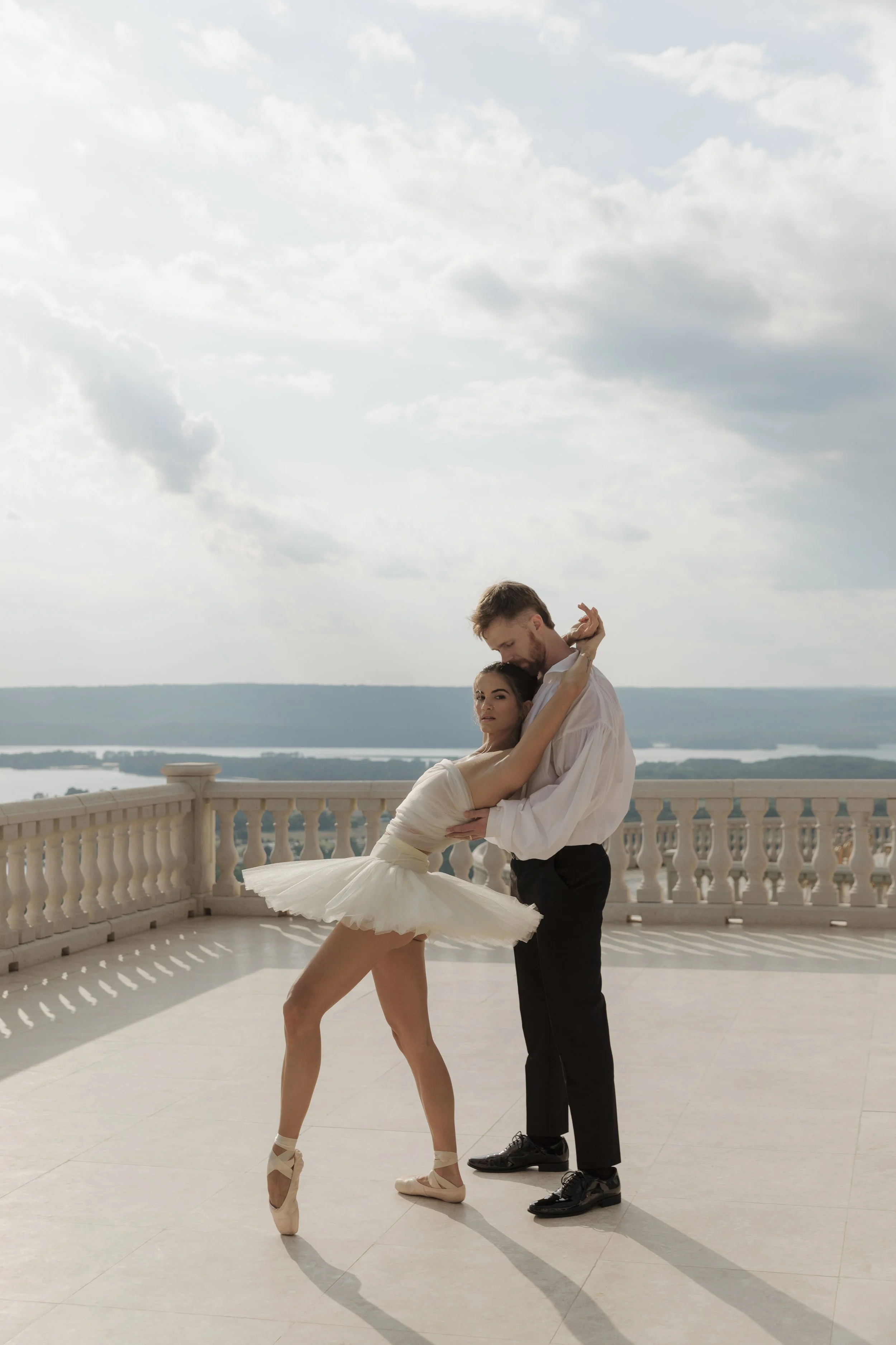 A man and a woman dancing on a balcony with a scenic view of water and hills in the background. The woman is in a white tutu and pointe shoes, and the man is in black pants and a white shirt.