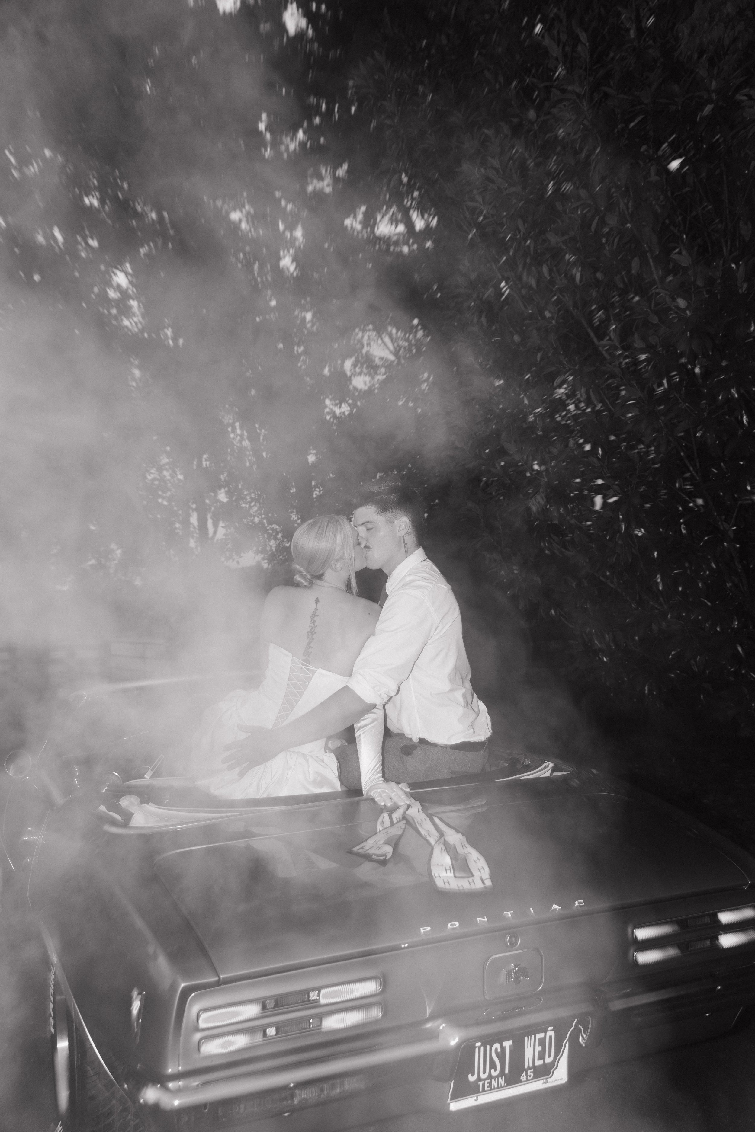 A black and white photo of a young couple kissing while sitting on the hood of a car in a wooded area. The woman is wearing a strapless dress, and the man is in a shirt. The car has a Tennessee license plate that reads "JUST WED".