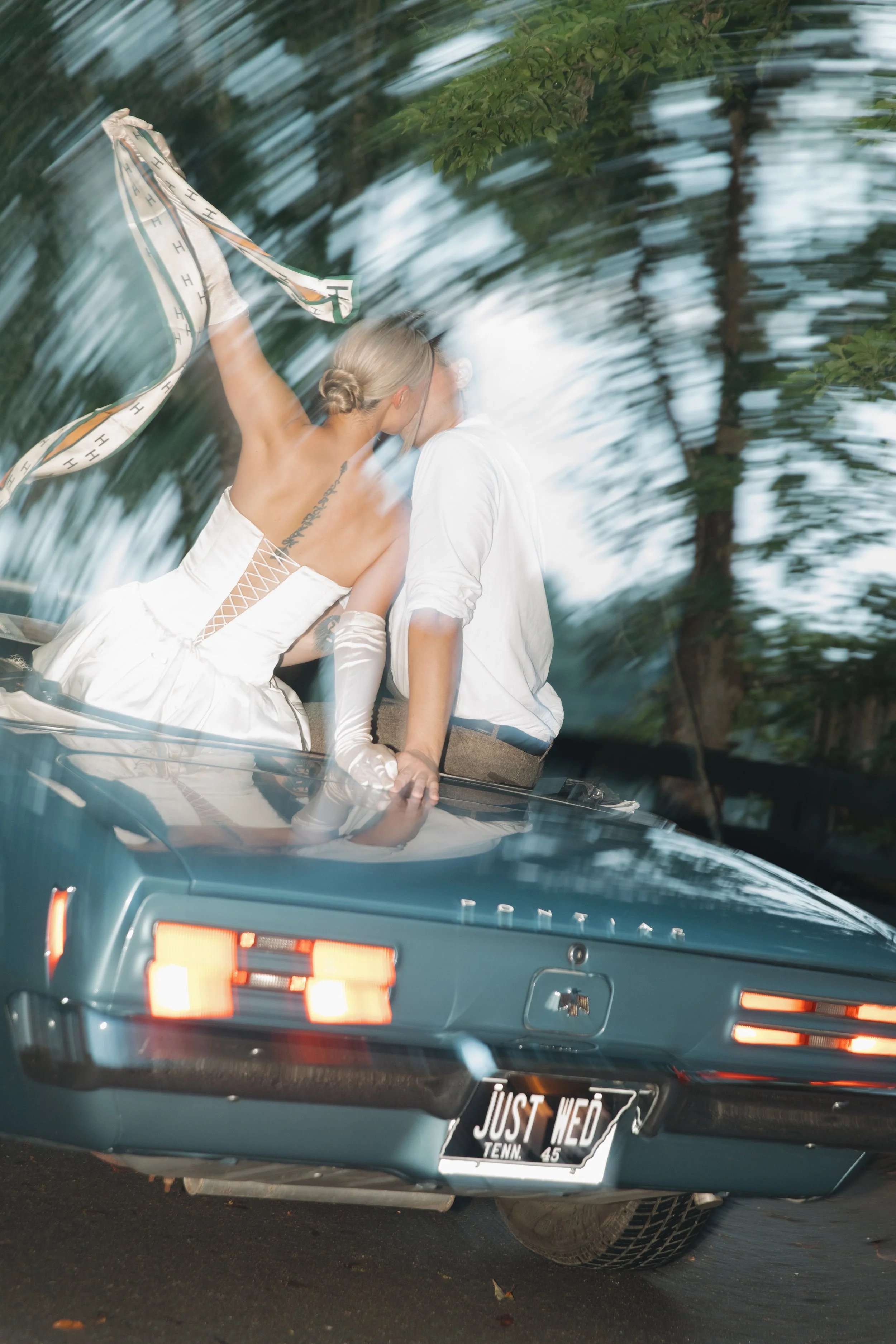 A couple is kissing in the back of a vintage blue car. The woman is wearing a white dress with gloves and a tattoo on her arm, holding a piece of cloth. The man is dressed in a white shirt. The license plate reads "JUST WED," indicating they are newly married. The photo is taken through the car's rear window, which creates a blurred, artistic effect.