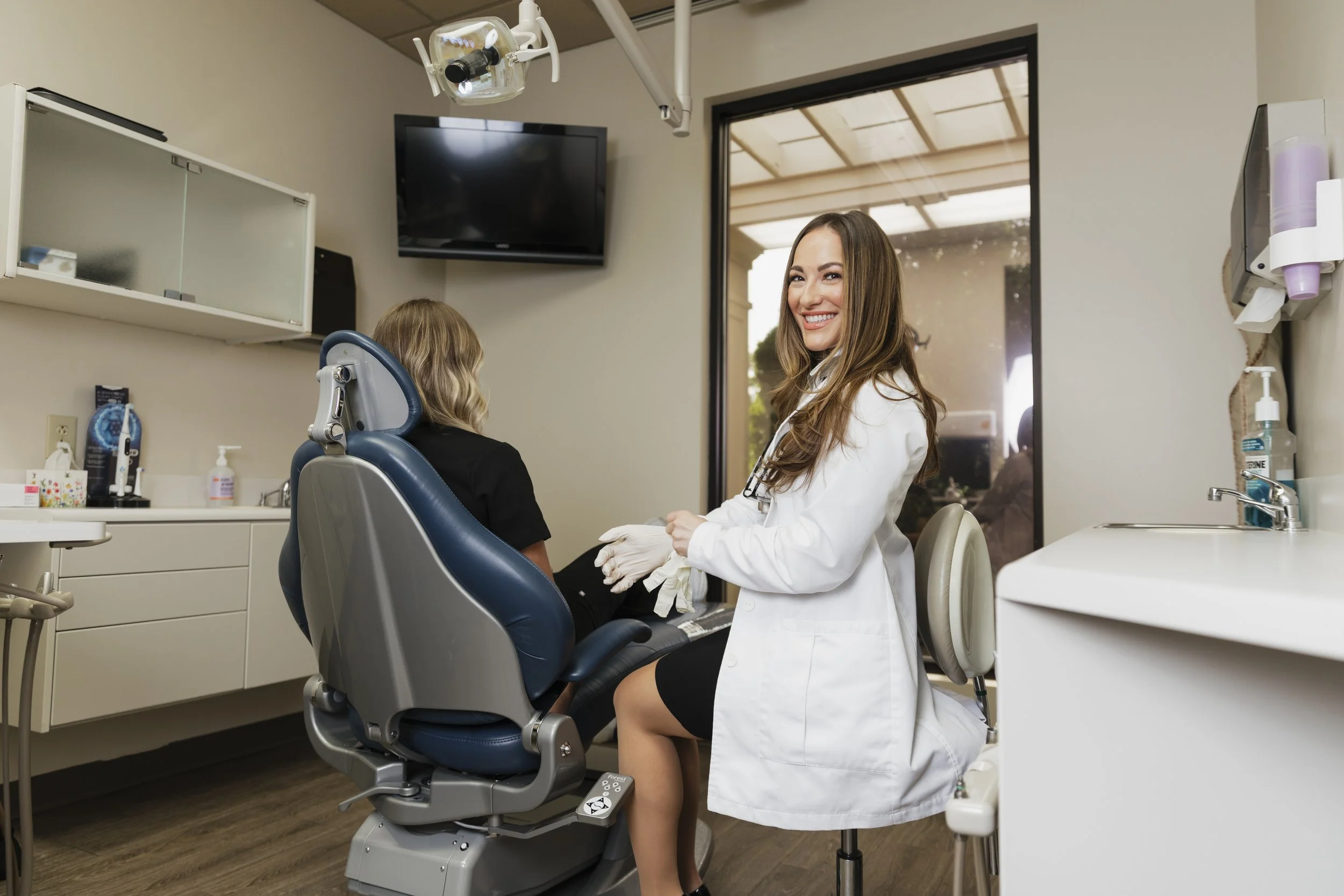 A female dentist with long brown hair smiling in a dental office, sitting next to a young female patient in a dental chair. The dental office has a window, a wall-mounted TV, and dental equipment visible.