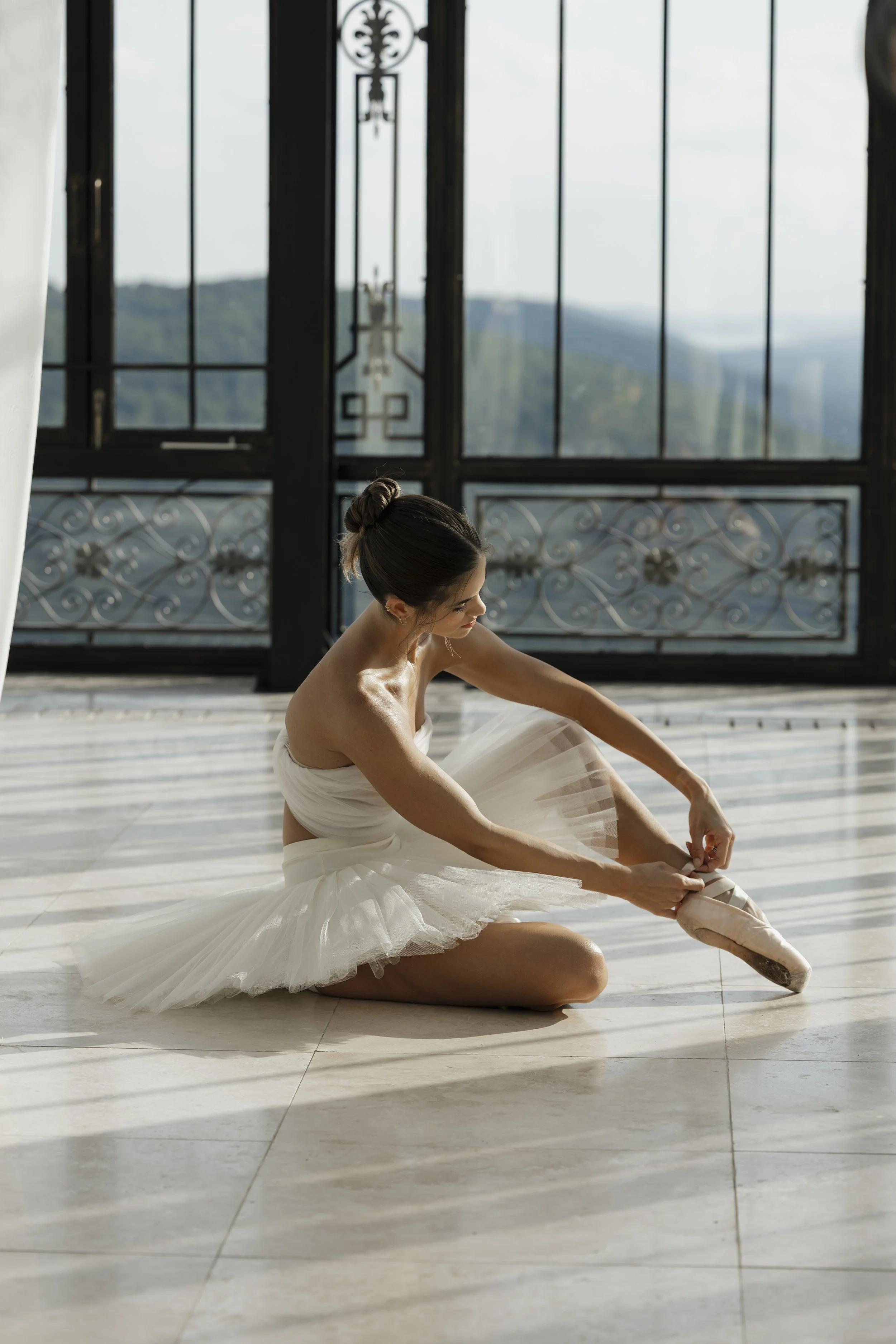 Bride seated on marble floor tying her satin pointe shoe ribbons in front of wrought iron and glass arched window with sheer drapery overlooking Alabama mountains, ballerina bridal editorial at Stone Haven Weddings by middle Tennessee wedding photogr