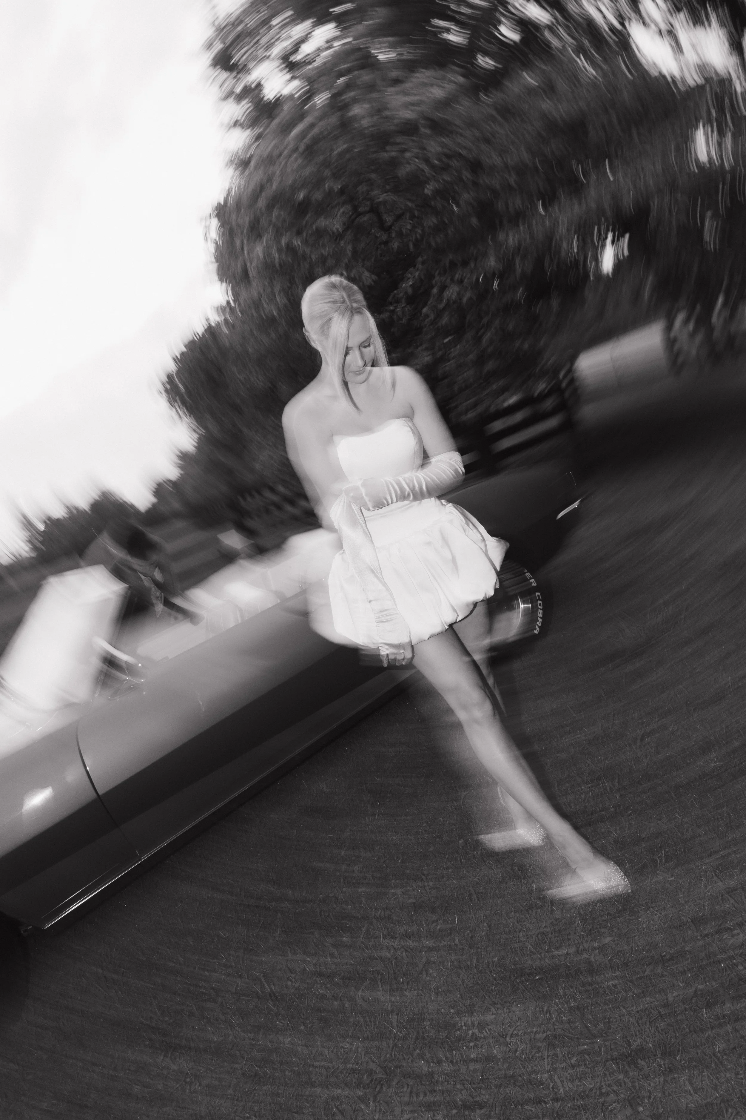 A woman dressed in a white strapless dress and long gloves sitting on a bench in a park, with a large tree and street lamps in the background. The photo has a blurry, spiral effect.