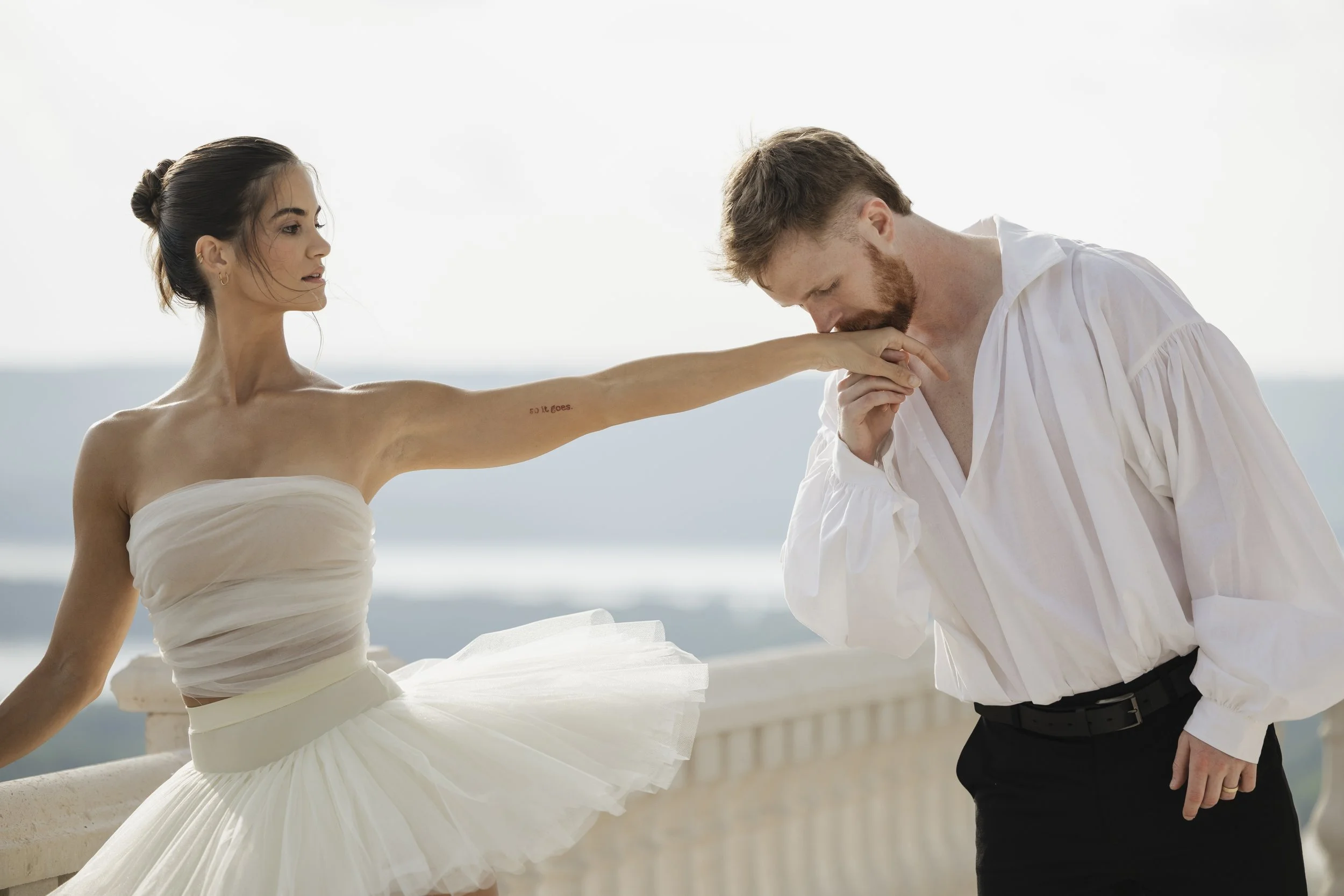 A woman in a strapless white dress extends her arm with a tattoo towards a man in a white shirt who is kissing her hand. They are outdoors with a cityscape and sky in the background.