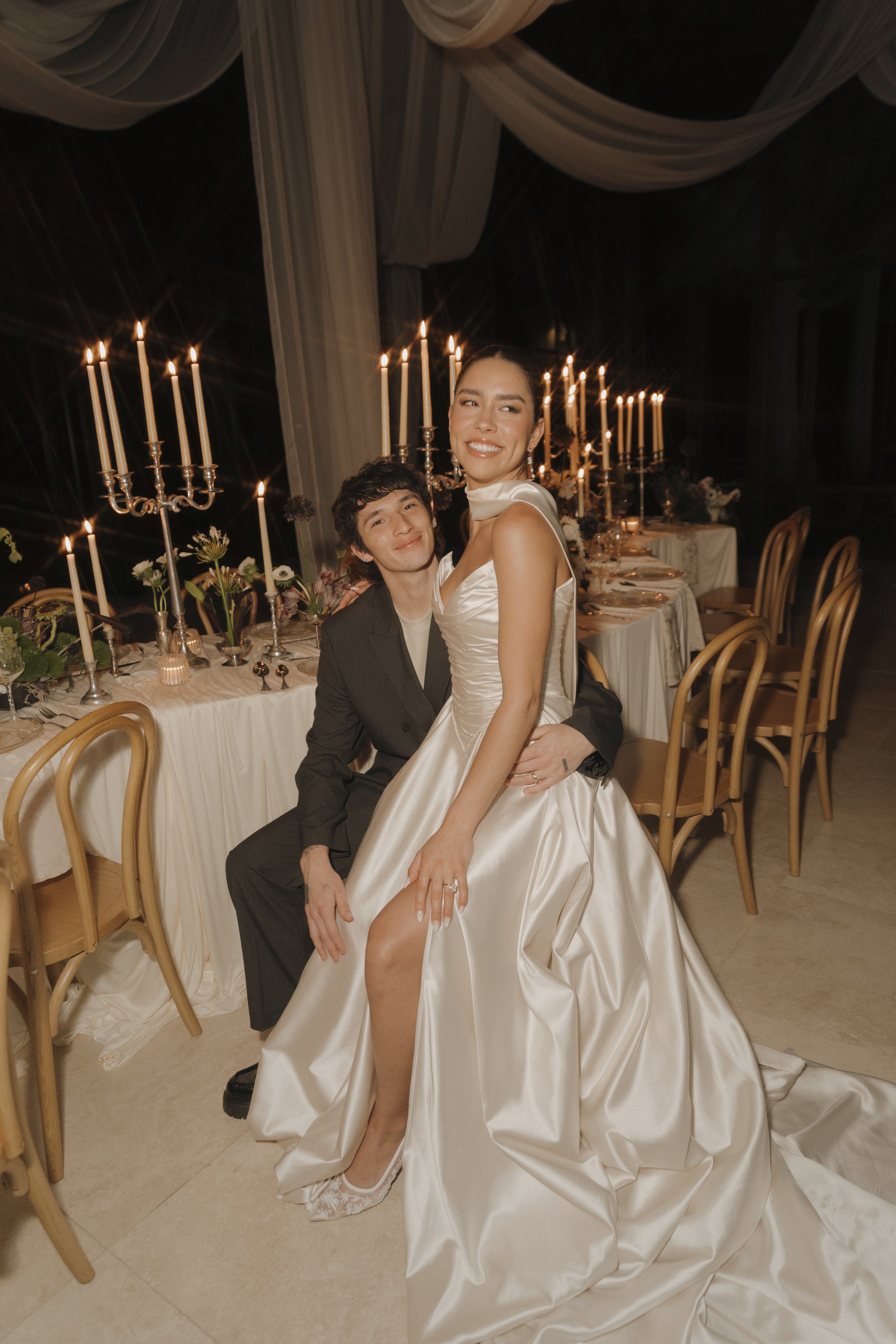 A woman in a wedding dress sitting on the lap of a man in a black suit at a formal event with a decorated table and candlelight in the background.