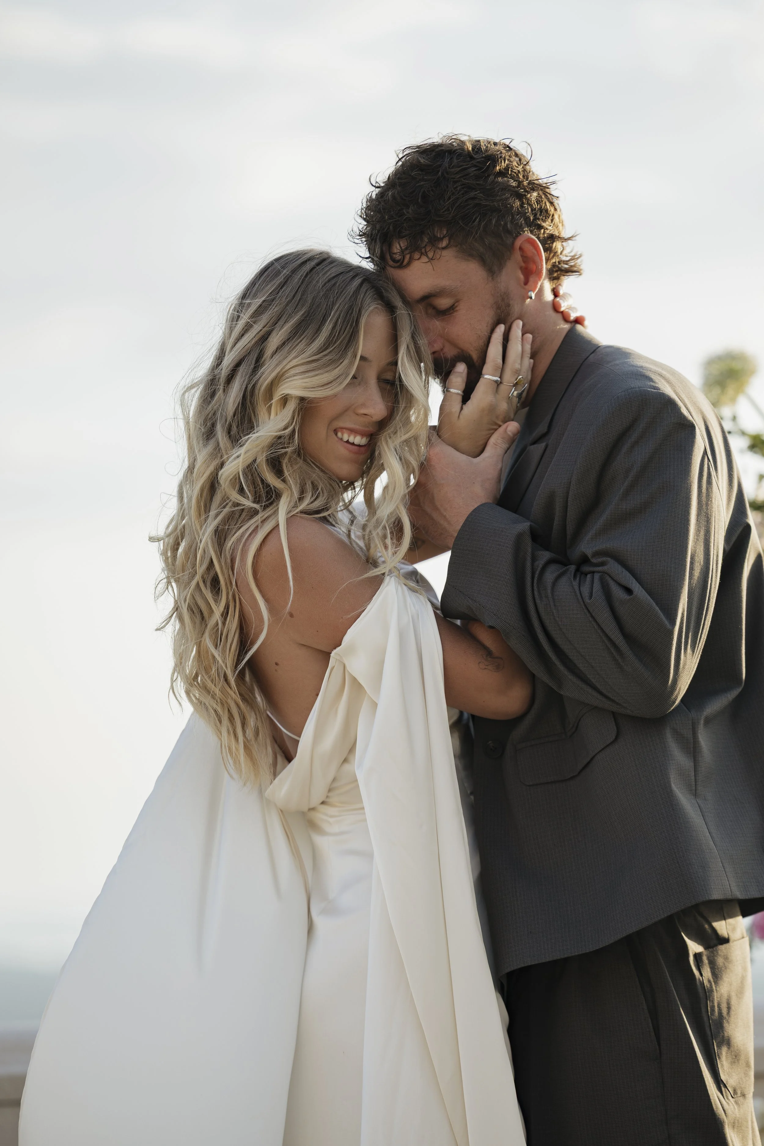 A woman and a man embrace tenderly outdoors during daylight, with eyes closed and smiling, featuring a cloudy sky background.