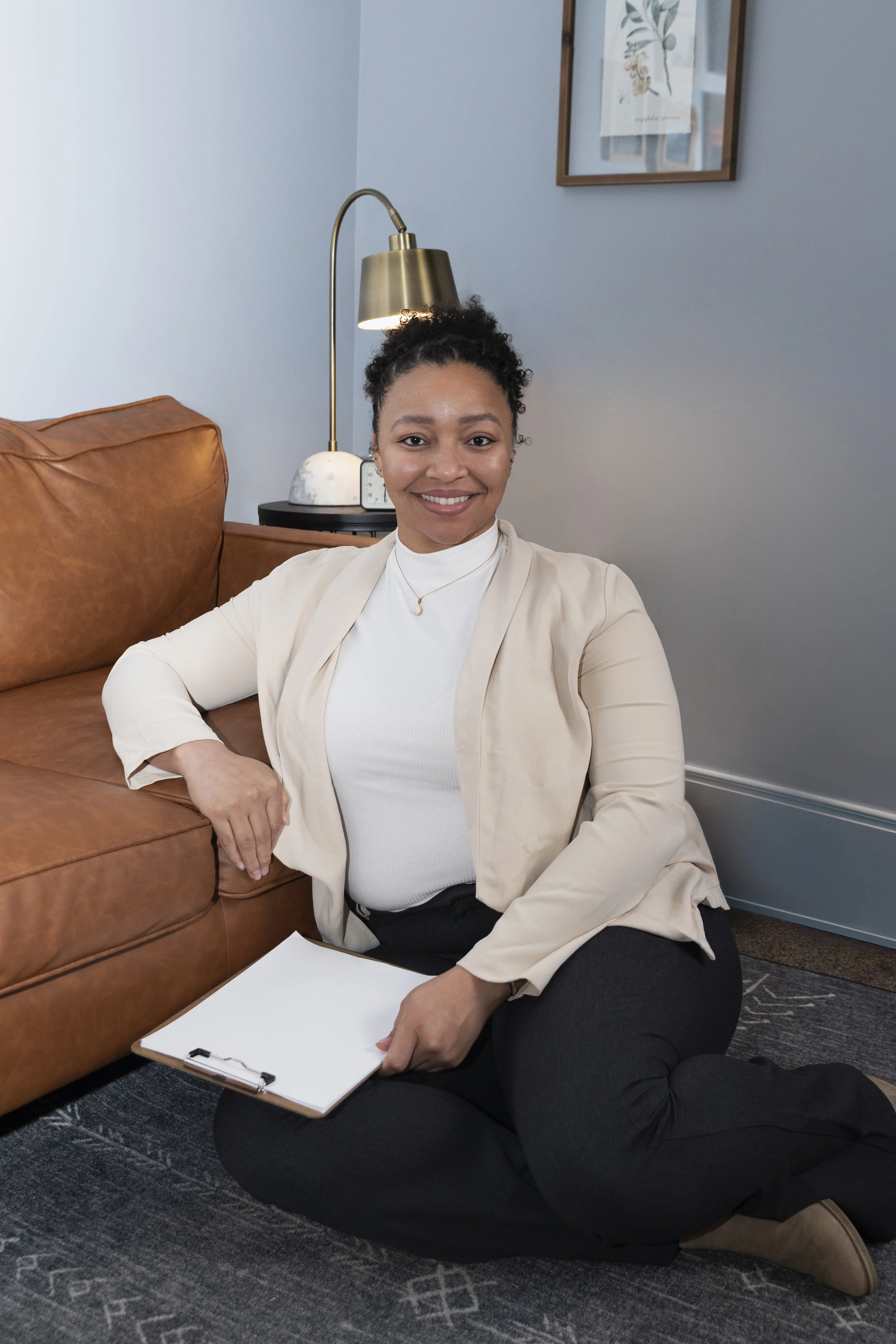 A woman sitting on the floor next to a brown leather couch, smiling at the camera, with a notepad on her lap, in a cozy room with a gray wall, a framed botanical print, and a table lamp in the background.