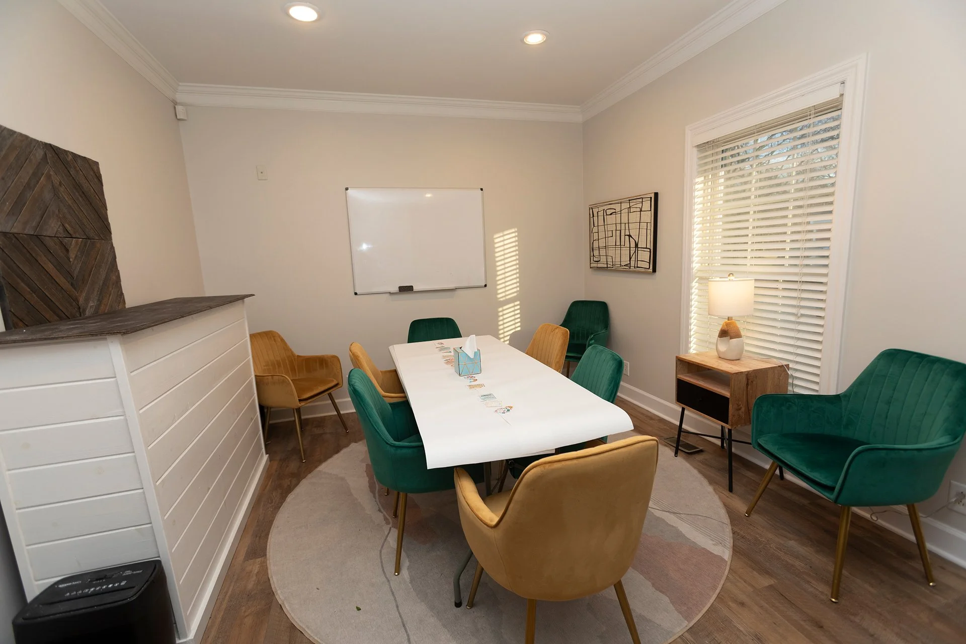 A cozy meeting room with a white rectangular table surrounded by cream and green velvet chairs. There is a large window with white blinds letting in natural light, a small wooden side table with a lamp, a decorative wall art piece, a whiteboard, and a tilted wooden panel on one wall.