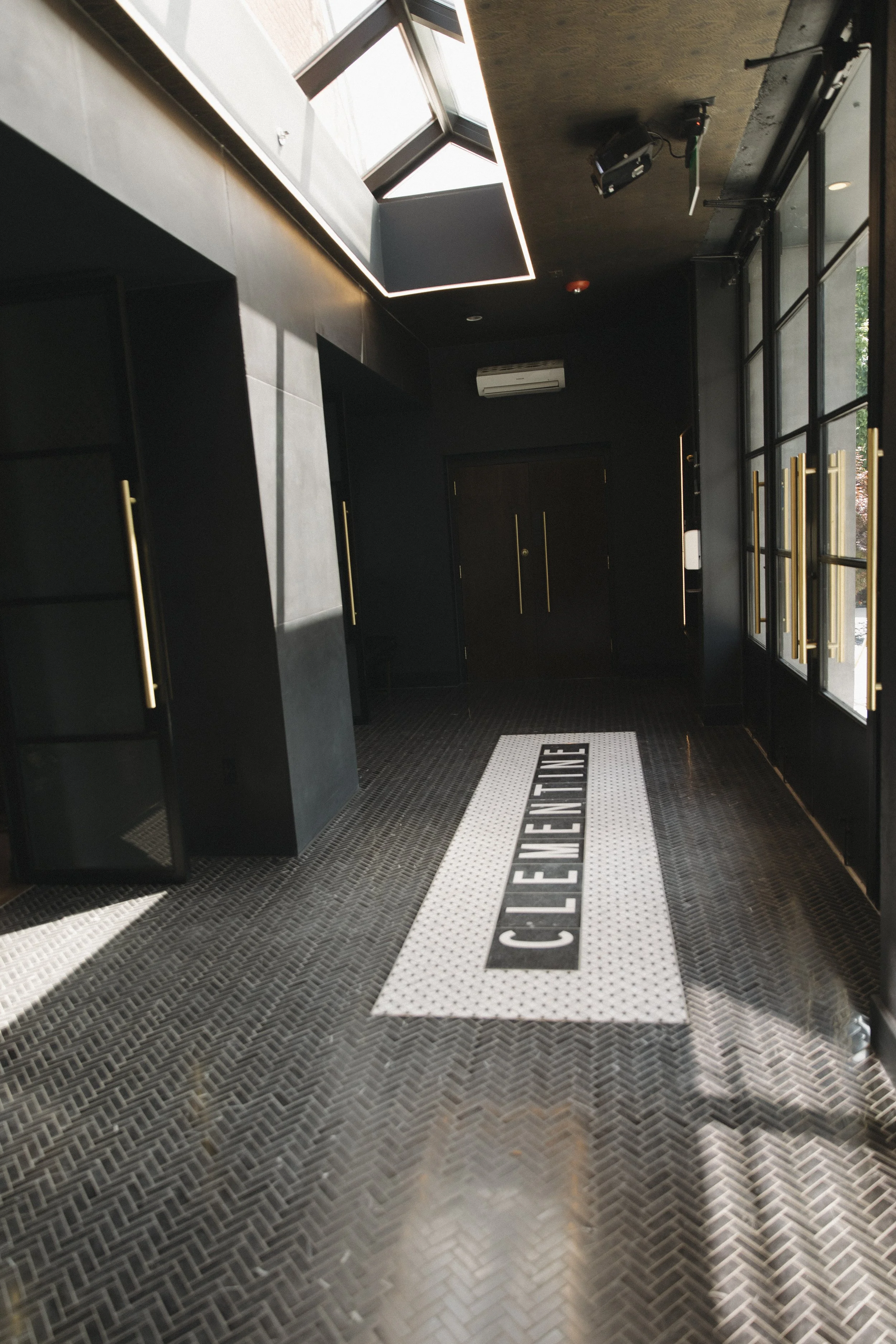The image shows a dark-colored entrance hallway with black lockers, black and white checkered flooring, and a white directional floor sticker that reads 'CIRCULATION.' There are large windows allowing natural light into the space.
