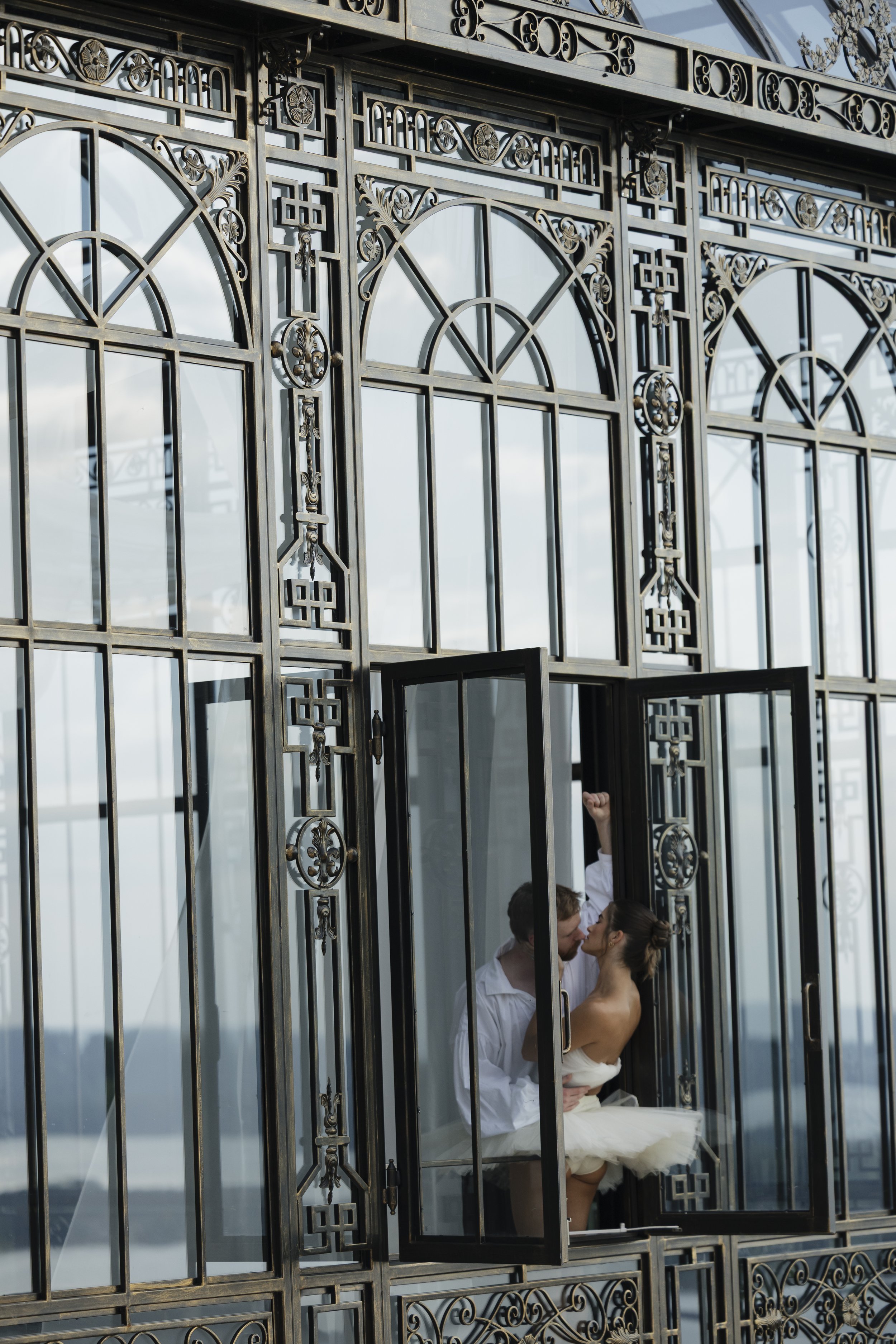 A couple sharing a kiss inside a decorative metal and glass structure, possibly a church or a chapel, with the man wearing a white shirt and the woman in a white dress. The structure has intricate metalwork and large windows, and a person is seen hol