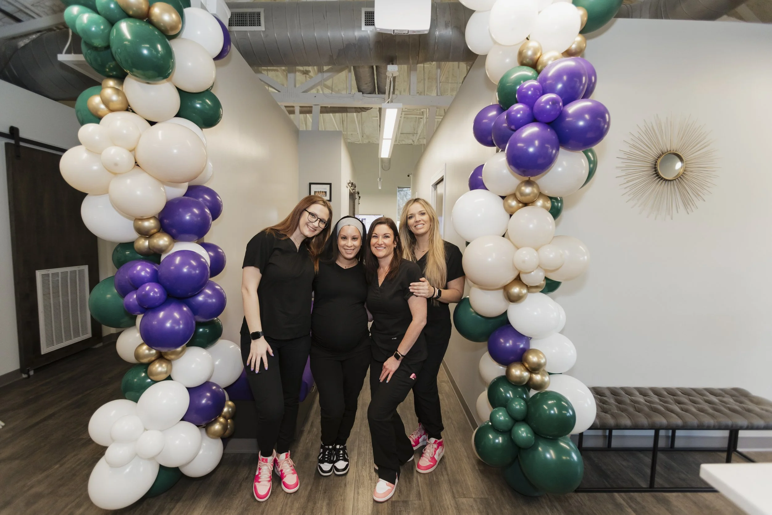 Four women standing inside a balloon arch at an indoor celebration, smiling and posing for the camera.