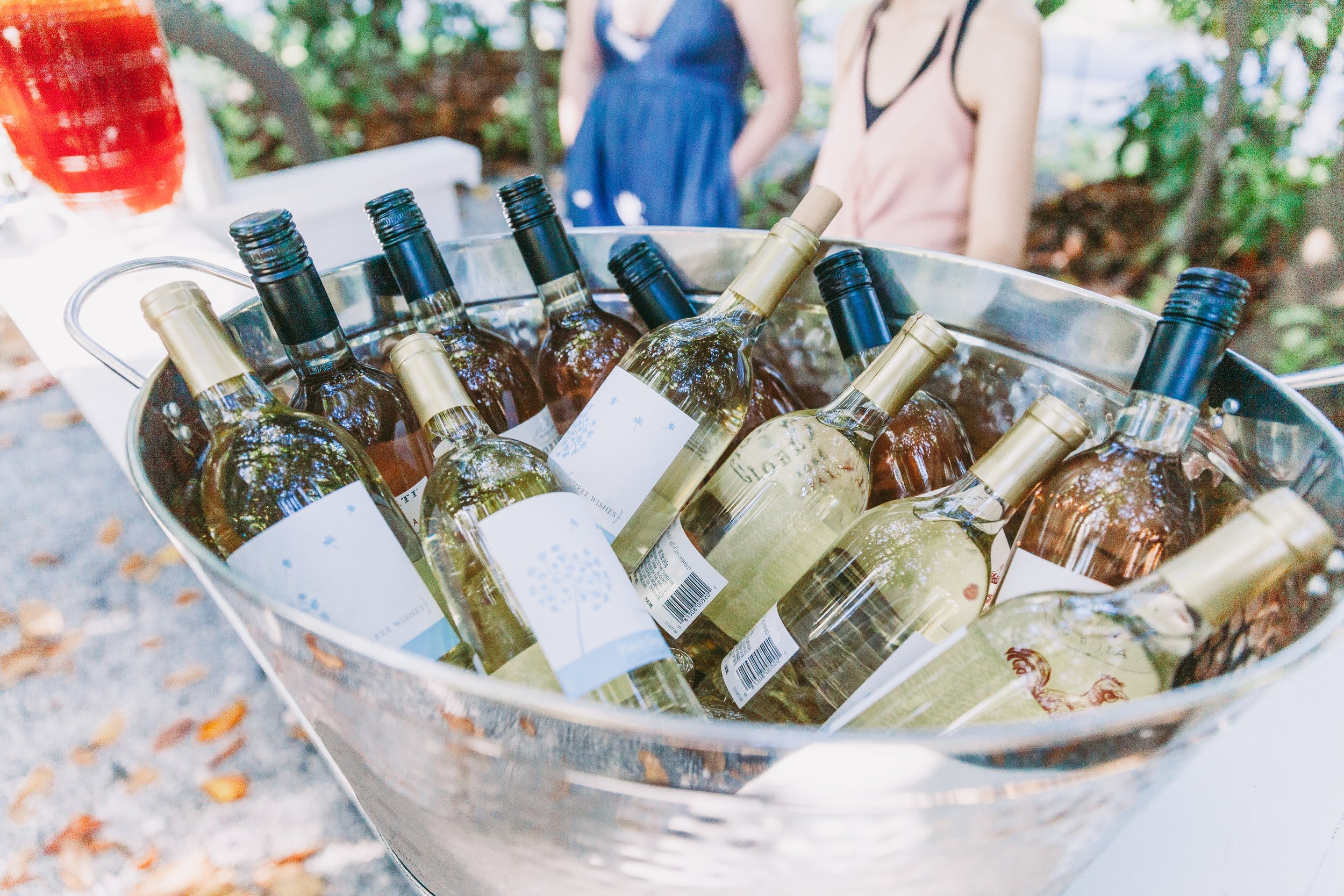 A metal bucket filled with bottles of white wine, with two people standing in the background outdoors.