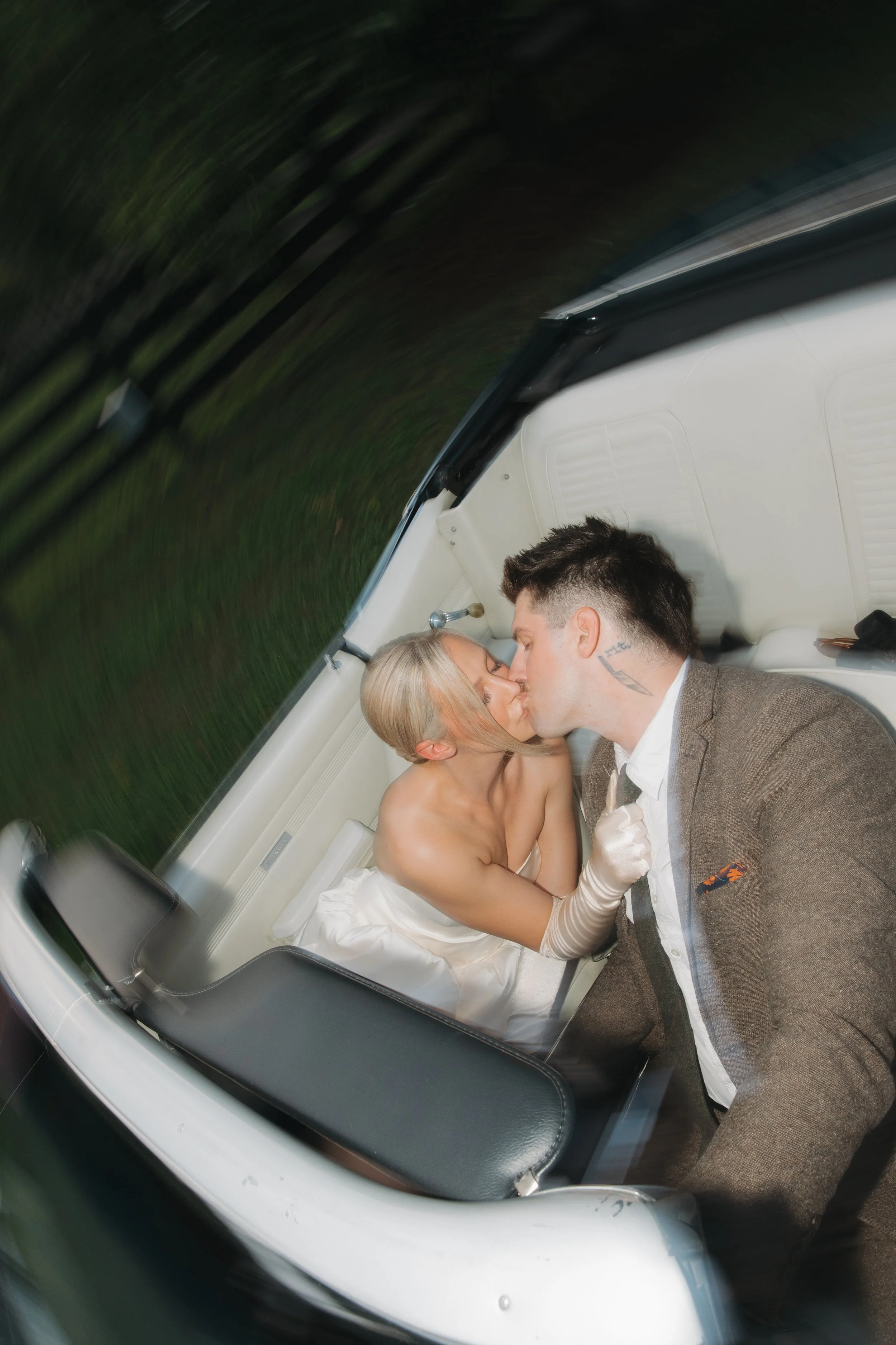 A woman and a man sharing a kiss inside a vintage car while the woman is wearing a strapless wedding dress and the man is dressed in a suit.