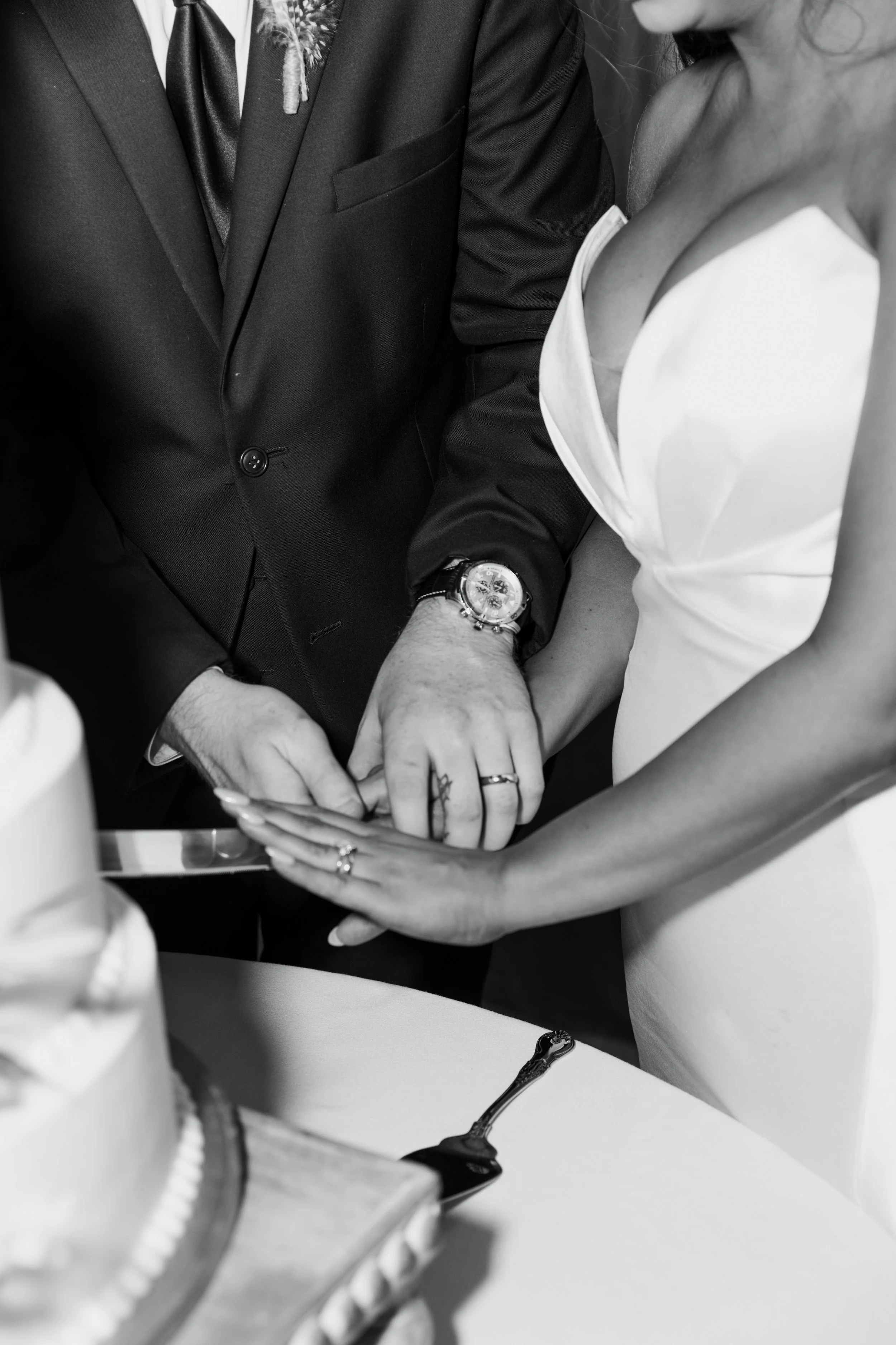 Close-up of a couple holding hands during a wedding ceremony, with both wearing wedding rings. The groom is dressed in a tuxedo, and the bride is in a white dress with a deep neckline.