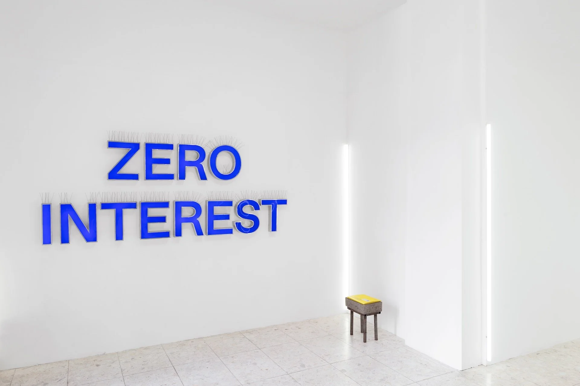 Blue lettering reading Zero Interest on a white gallery wall above a small wooden stool