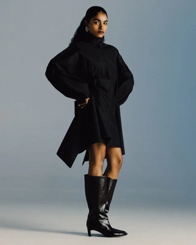 Black draped coat dress worn with knee-high black leather boots against a pale blue backdrop