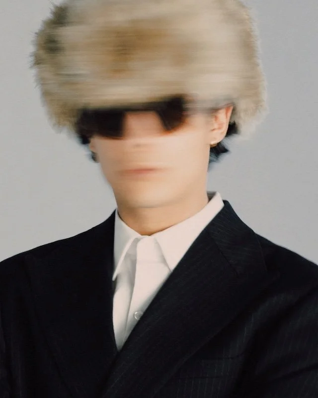 Cream faux fur hat pulled low over dark sunglasses, worn with a pinstripe suit and white shirt