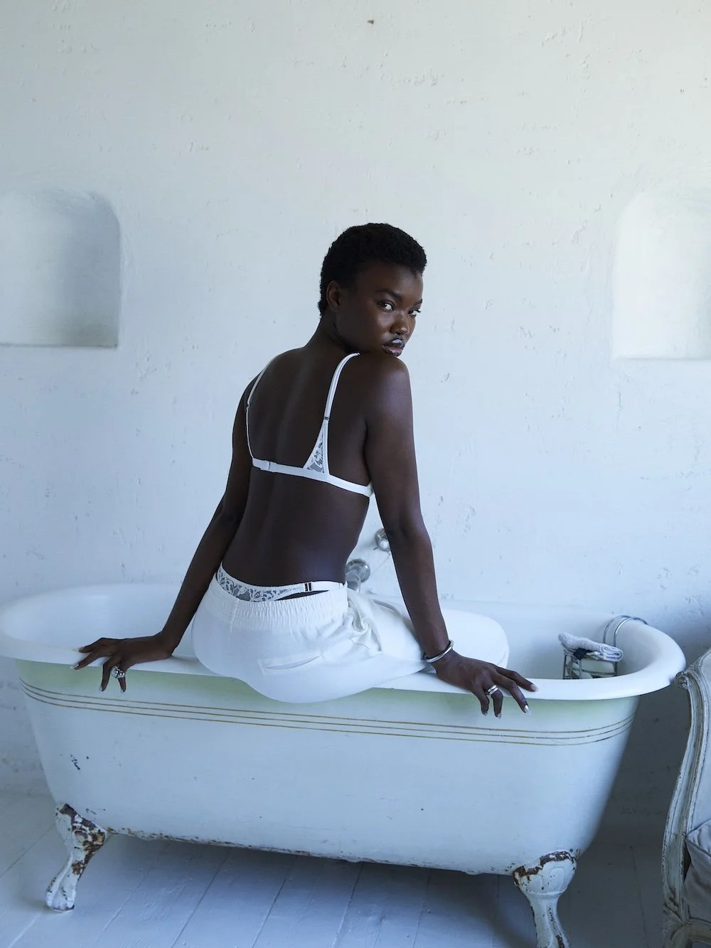 Model in Camila Martin Barla white lace lingerie and trousers, seated in a vintage bathtub in a minimal, white-washed space.