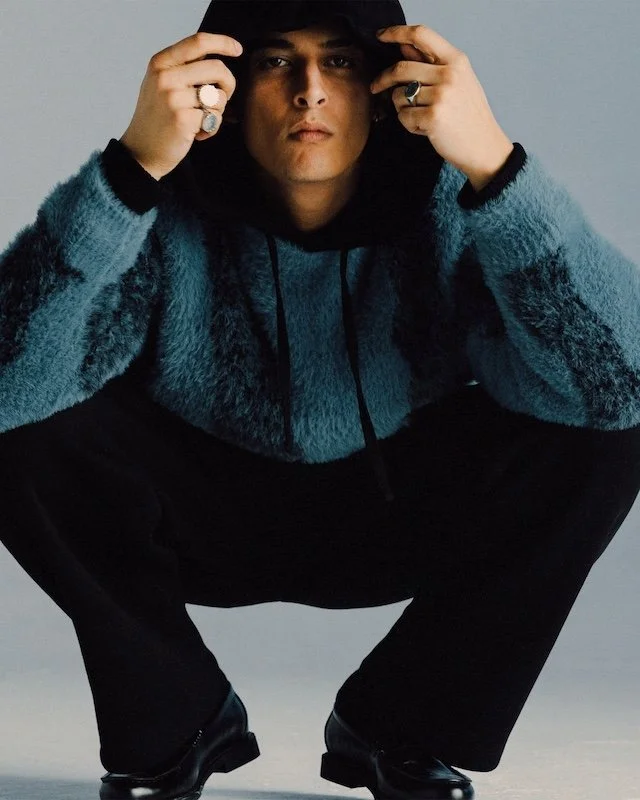 Model crouching, pulling up the hood of a teal brushed mohair jacket worn over black