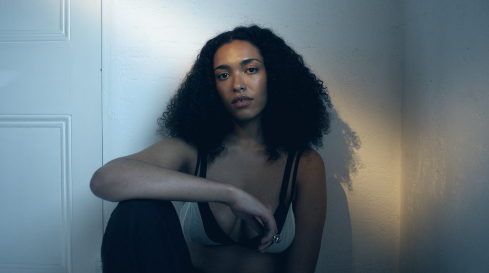 Model seated against a pale blue panelled wall in a white lingerie top and dark trousers