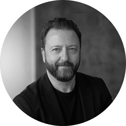 Black and white portrait of a man with a beard and short hair, wearing a dark blazer and t-shirt, smiling slightly, in front of a blurred background.