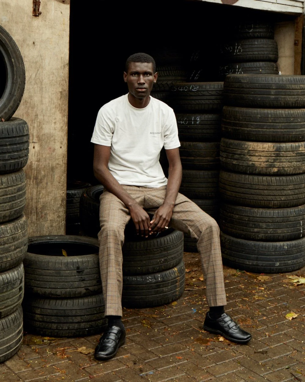 Model seated on stacked car tyres in a yard, wearing a cream logo T-shirt and beige checked trousers