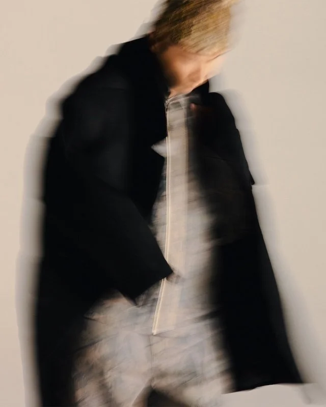 Black overcoat over pale grey trousers, blurred mid-walk against a white backdrop