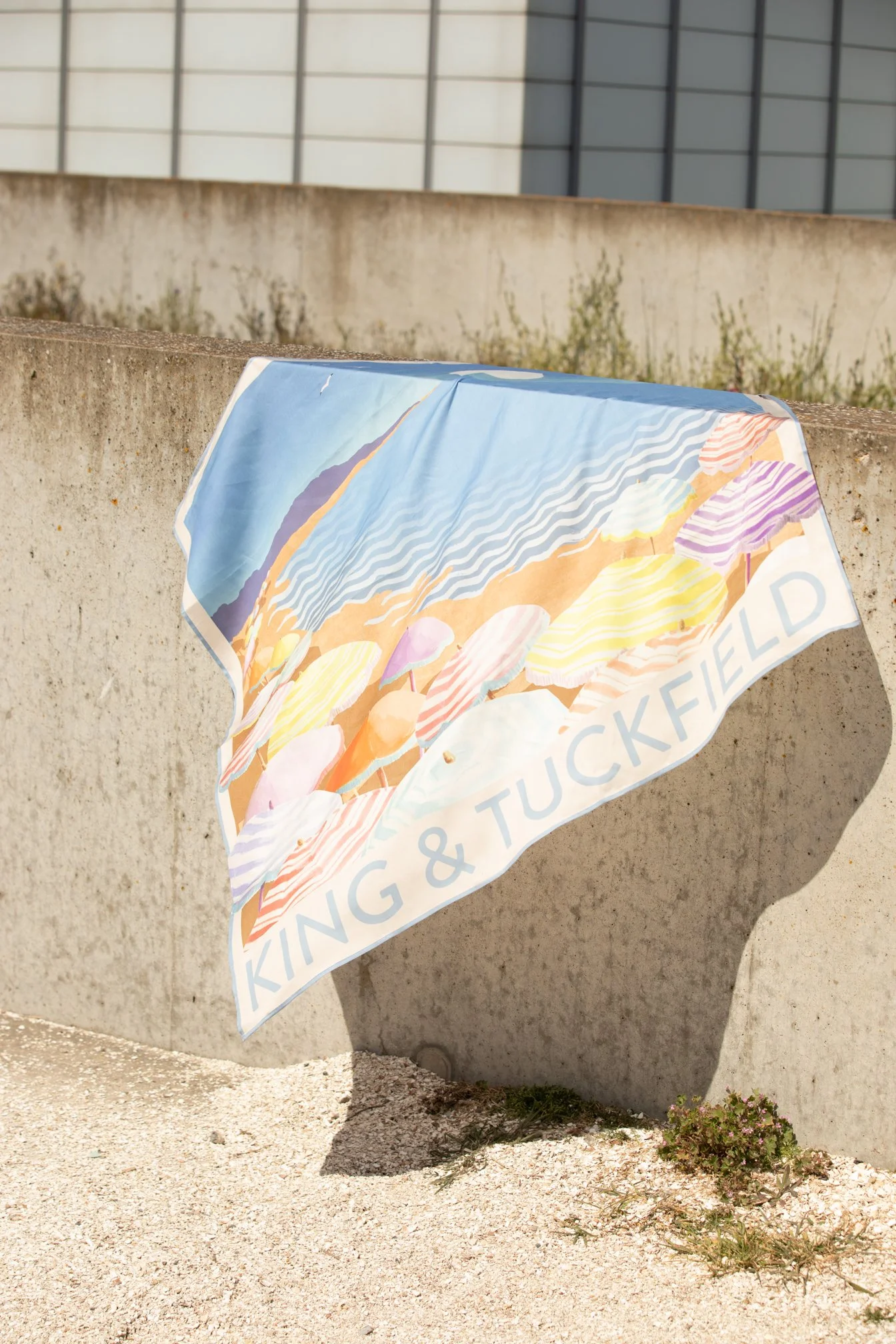 Printed silk scarf draped over a concrete wall, showing an illustrated beach scene and the King and Tuckfield name