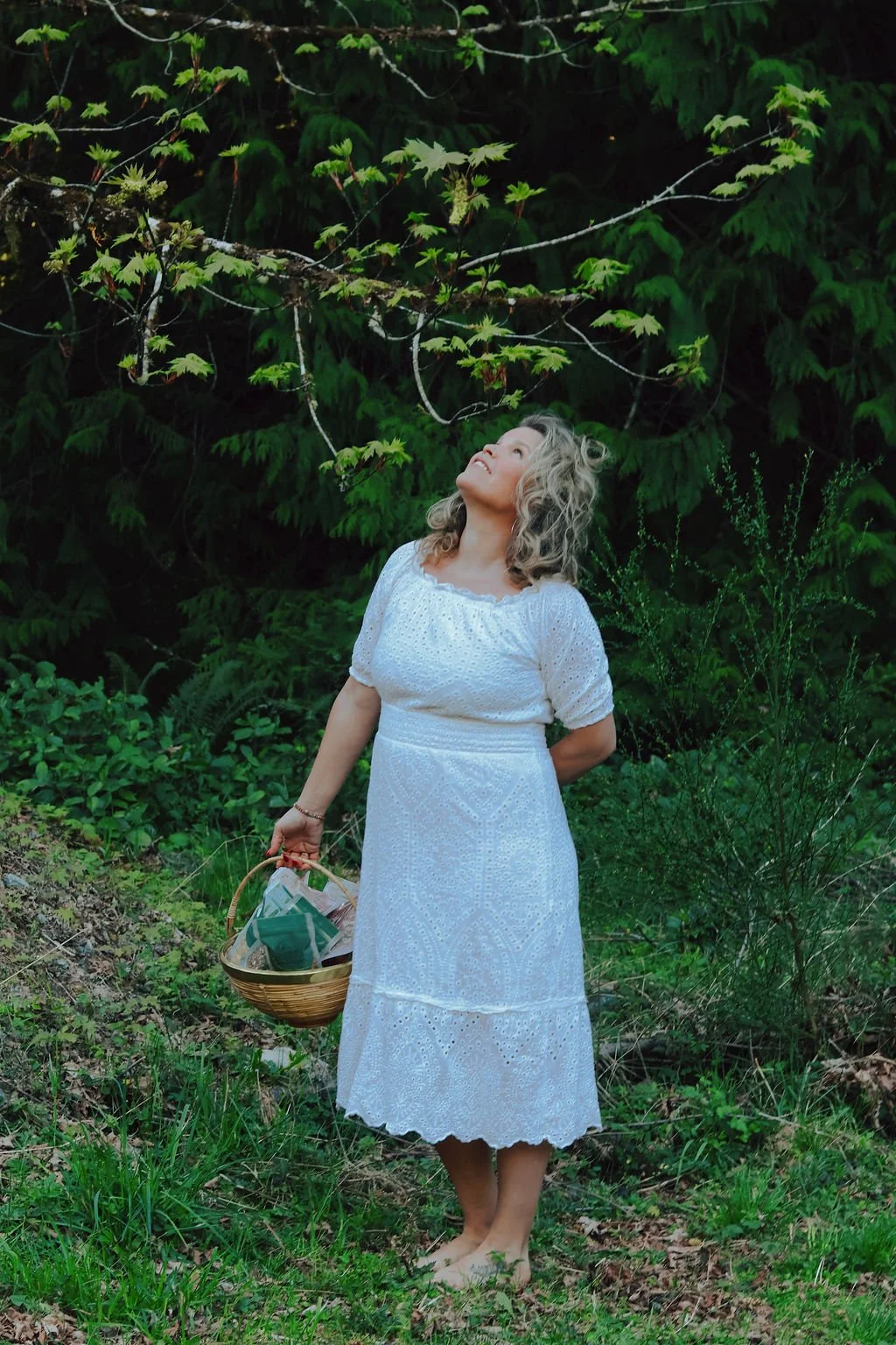 A woman in a white dress stands barefoot on grass in a forested area, holding a basket of Kisses To Go Organic Tea, looking up at the leaves on a tree branch.