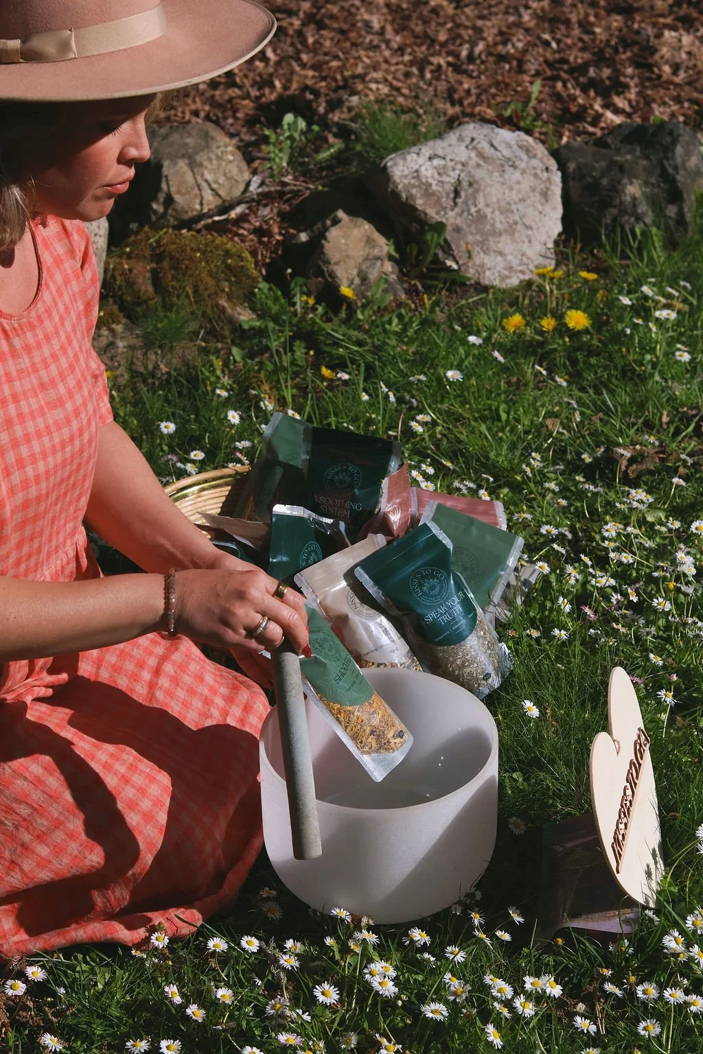 A woman in a peach checkered dress and wide-brimmed hat sound heals herbal tea outdoors in a meadow with daisies, surrounded by tea packages and crystal singing bowl.