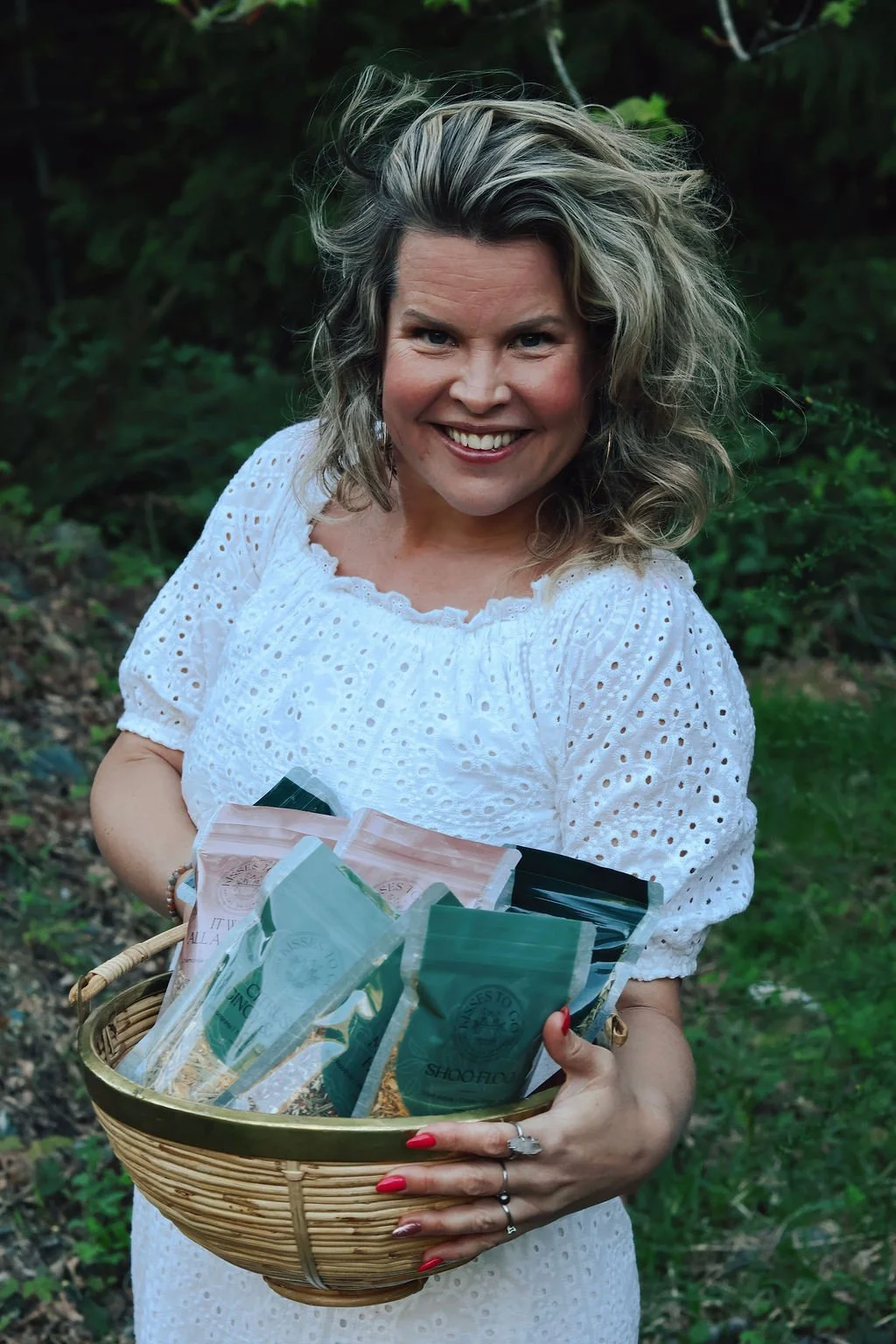 A smiling woman with curly, blonde hair holding a basket filled with various Kisses To Go artisan tea co tea blends