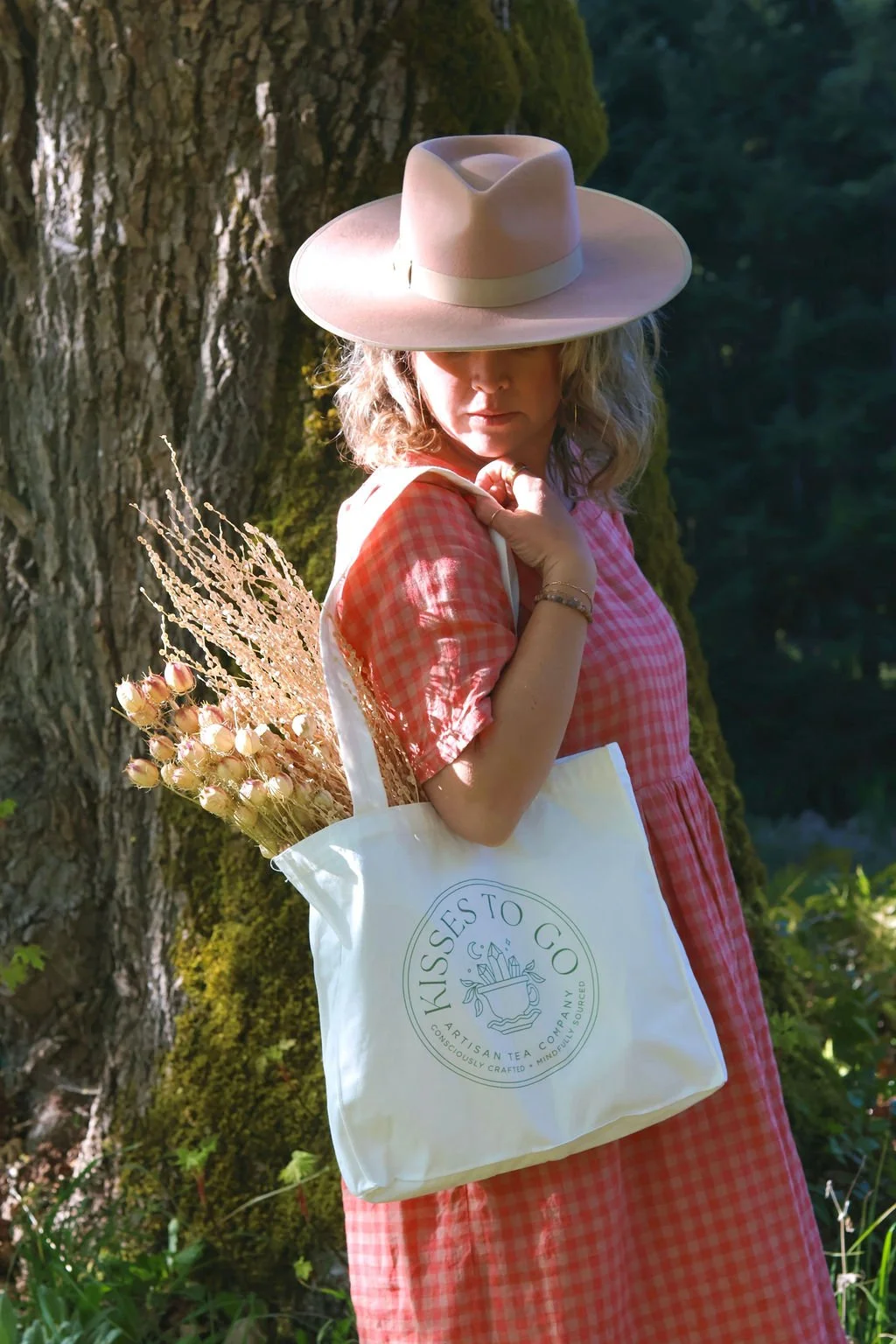 A woman with curly blonde hair wearing a wide-brimmed pink hat and a pink checkered dress, holding a white tote bag with a circular logo that reads 'kisses to  go Artisan Tea Company.'