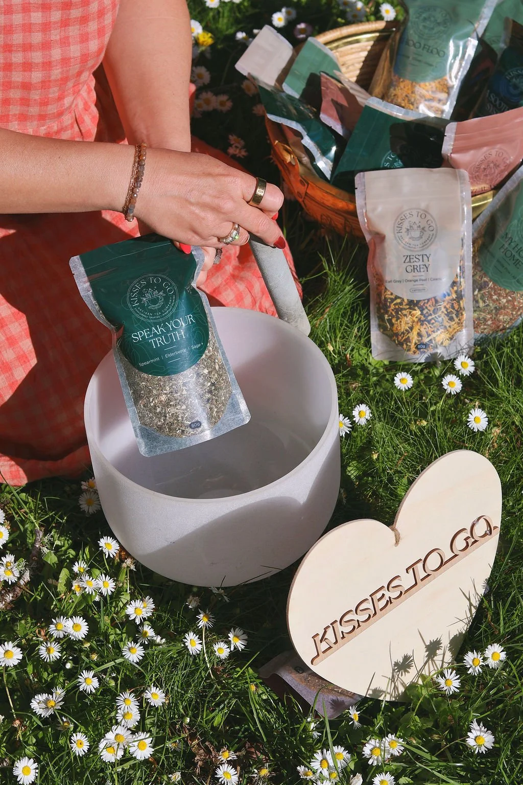A person is pouring loose herbal tea labeled 'Speak Your Truth' into crystal singing bowl. The scene is outdoors on grass with small daisies, with other herbal tea packages and a heart-shaped sign that says 'Kisses to Go'.