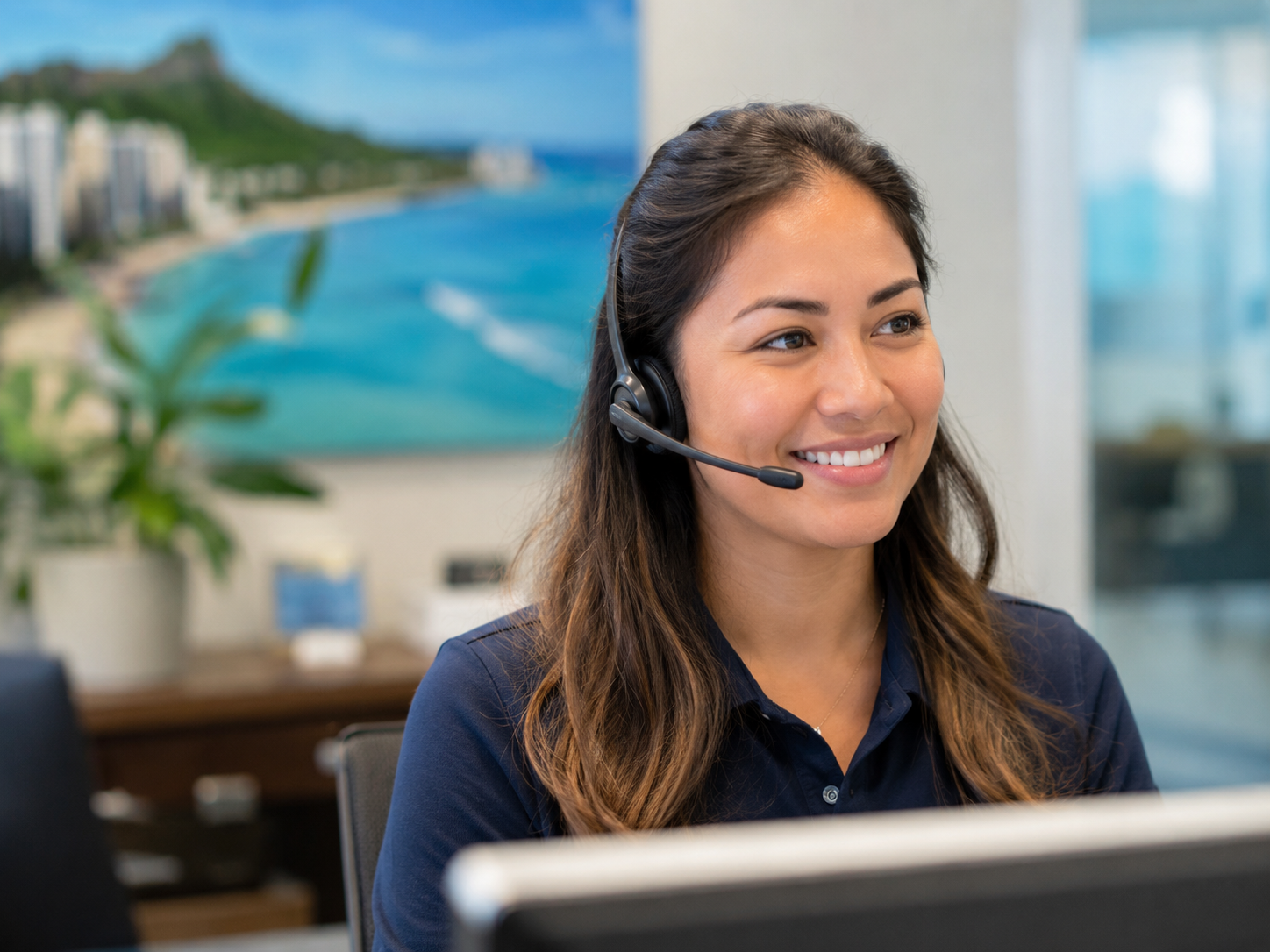 A woman wearing a headset smiles while working at a desk with a computer in an office with a beach and cityscape mural on the wall in the background.
