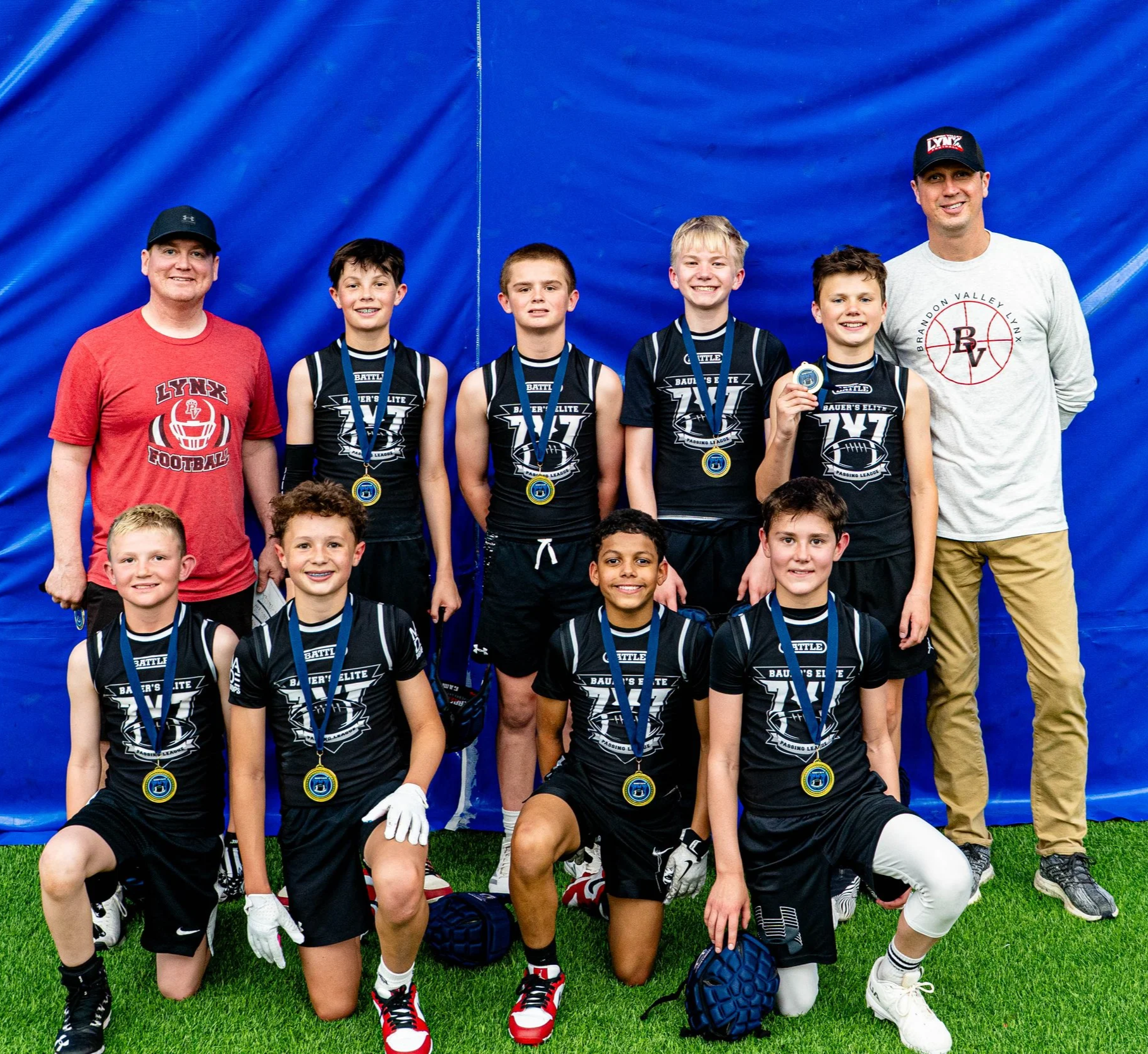 Youth football team with coaches, wearing black uniforms with medals, posing for a group photo in front of a blue background.
